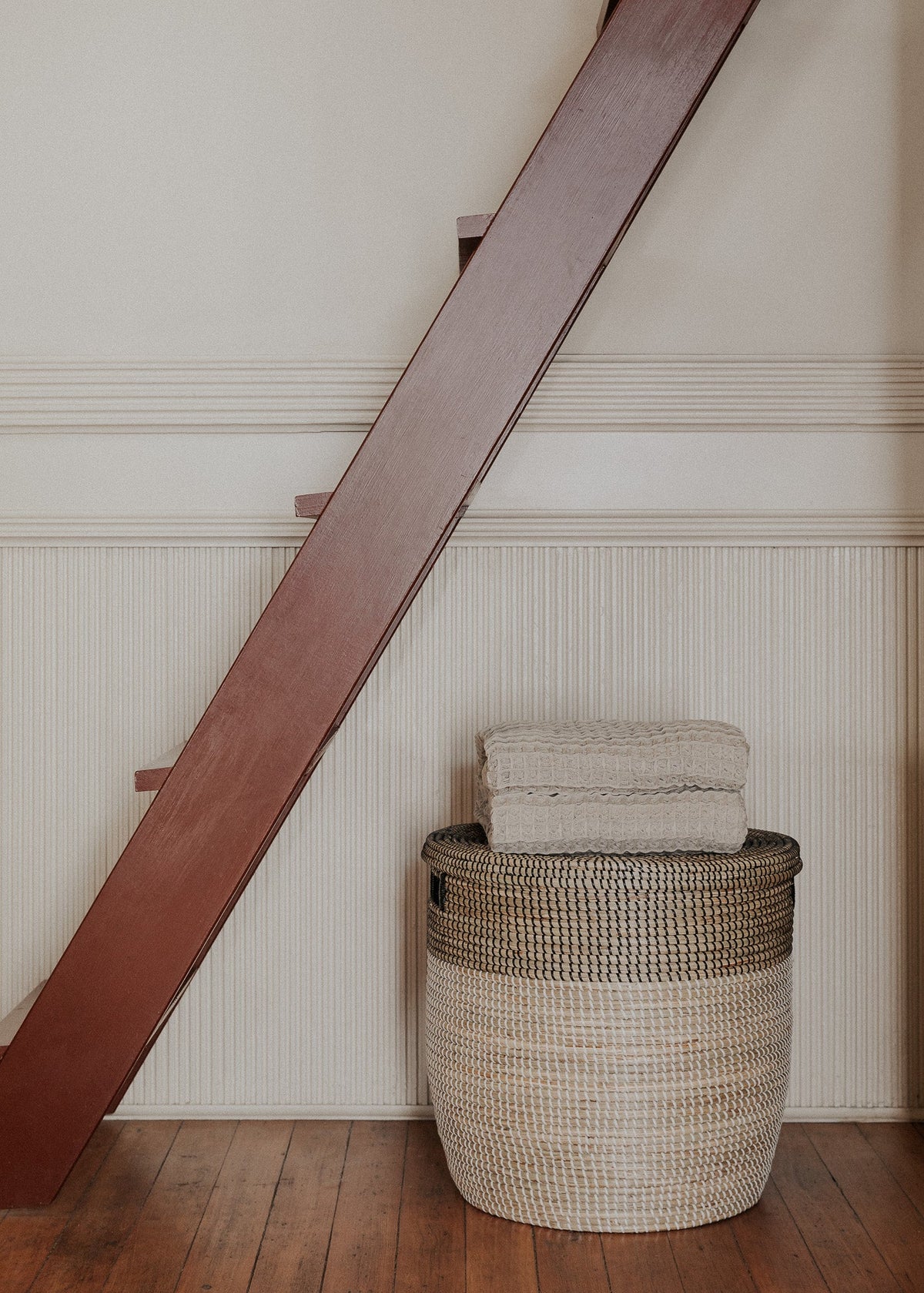 Large lidded basket styled beneath a wooden staircase with folded neutral textiles on top.