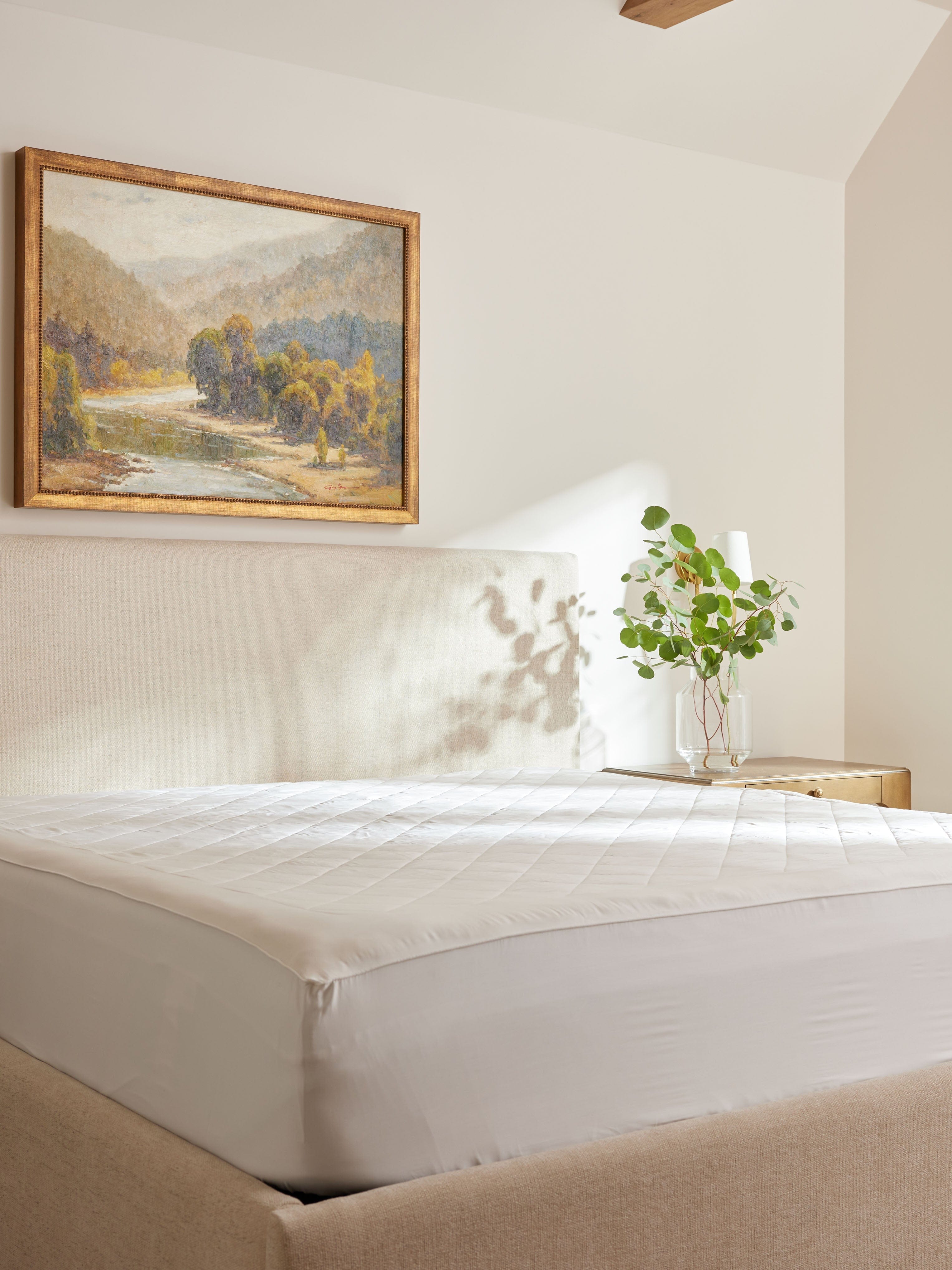 White mattress pad covering a mattress in a sunlit bedroom, with its fitted skirt extending down the sides beneath a landscape painting.