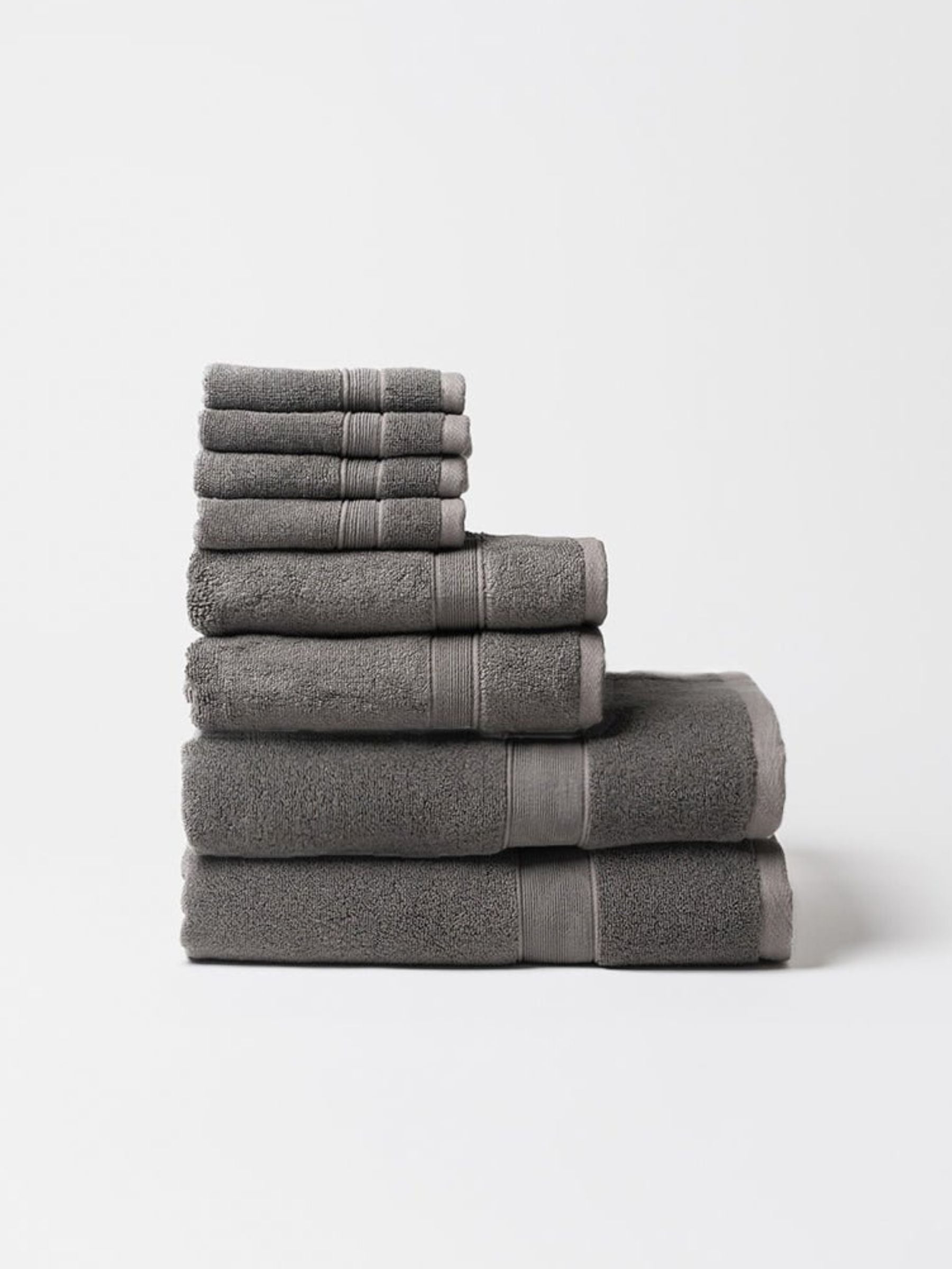 Charcoal gray towel set stacked by size on a pale background, showing thick folded edges, plush looped fabric, and matching woven border bands.