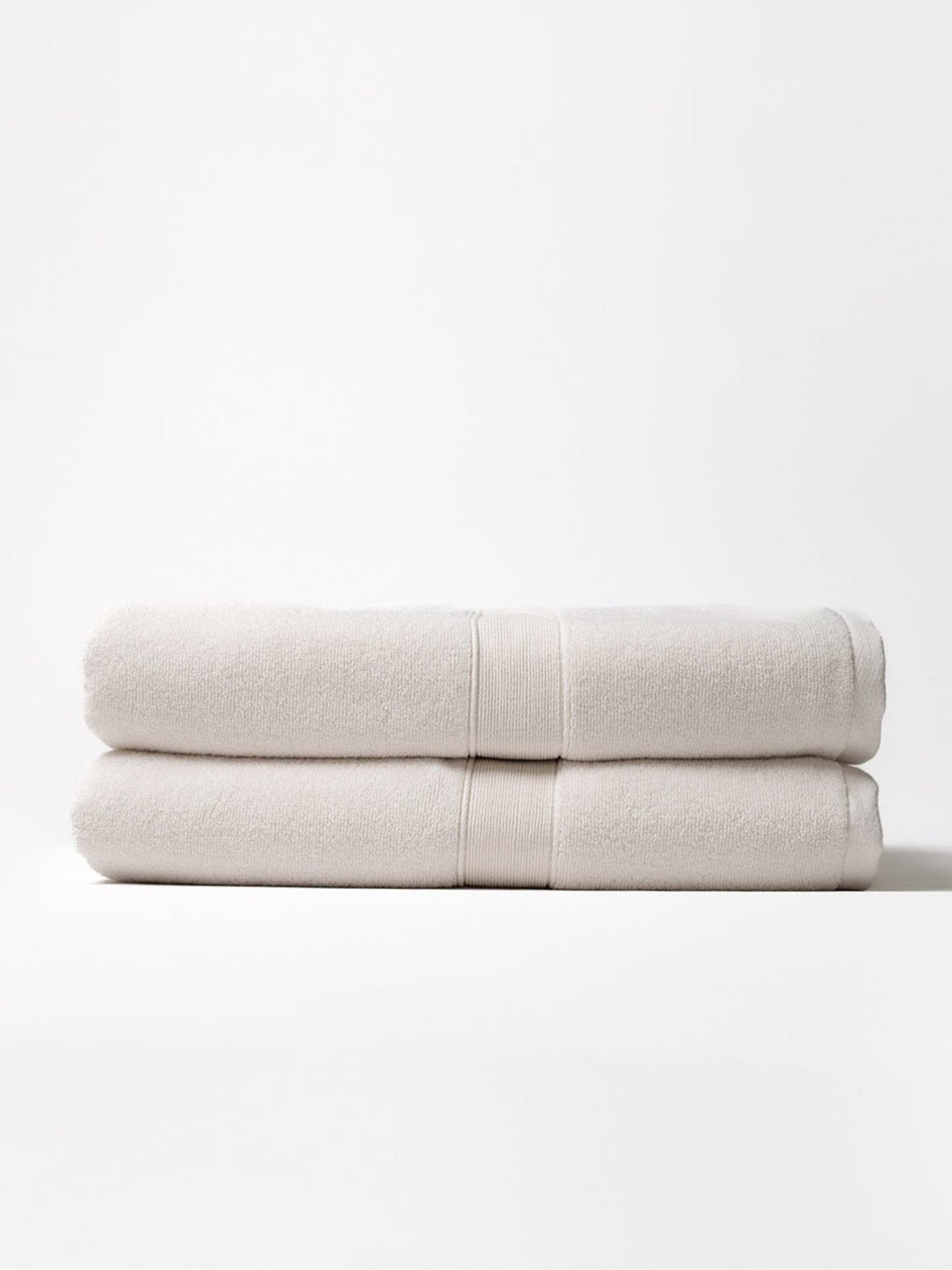 Two ivory Luxe Bath Sheets folded and stacked on a pale background, showing plush looped texture, thick folded edges, and matching woven border bands.