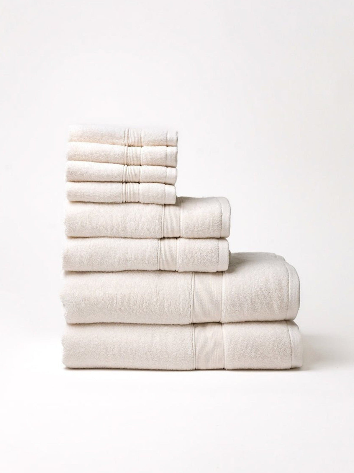 Ivory towel set stacked by size on a pale background, with four washcloths above two hand towels and two bath towels, showing plush texture and woven border bands.