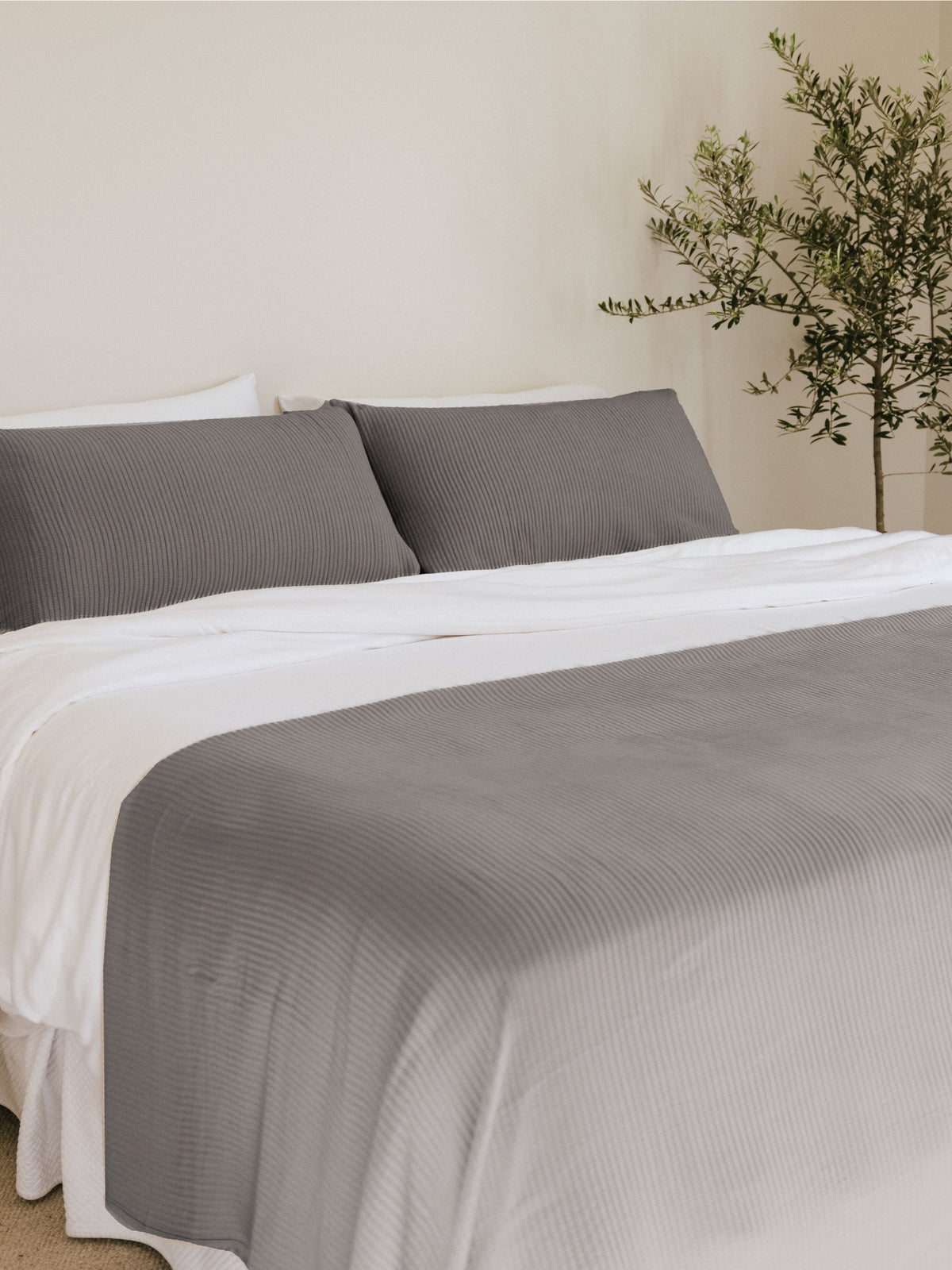 Gray coverlet draped across a bed with white sheets and matching gray pillows, showing its textured surface and rounded folds beside a leafy plant.