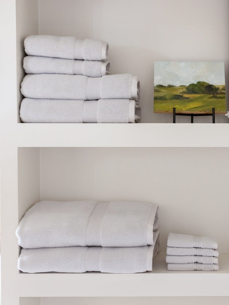 Light gray towels arranged on two white shelves beside a small landscape painting, with larger folded towels and a separate washcloth stack below.