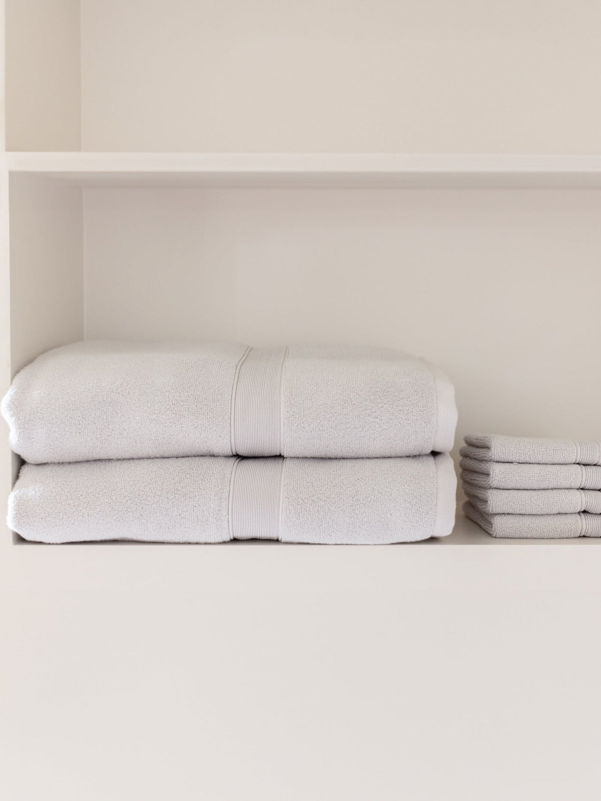 Light gray bath sheets stacked on a white bathroom shelf beside a separate pile of smaller folded towels.