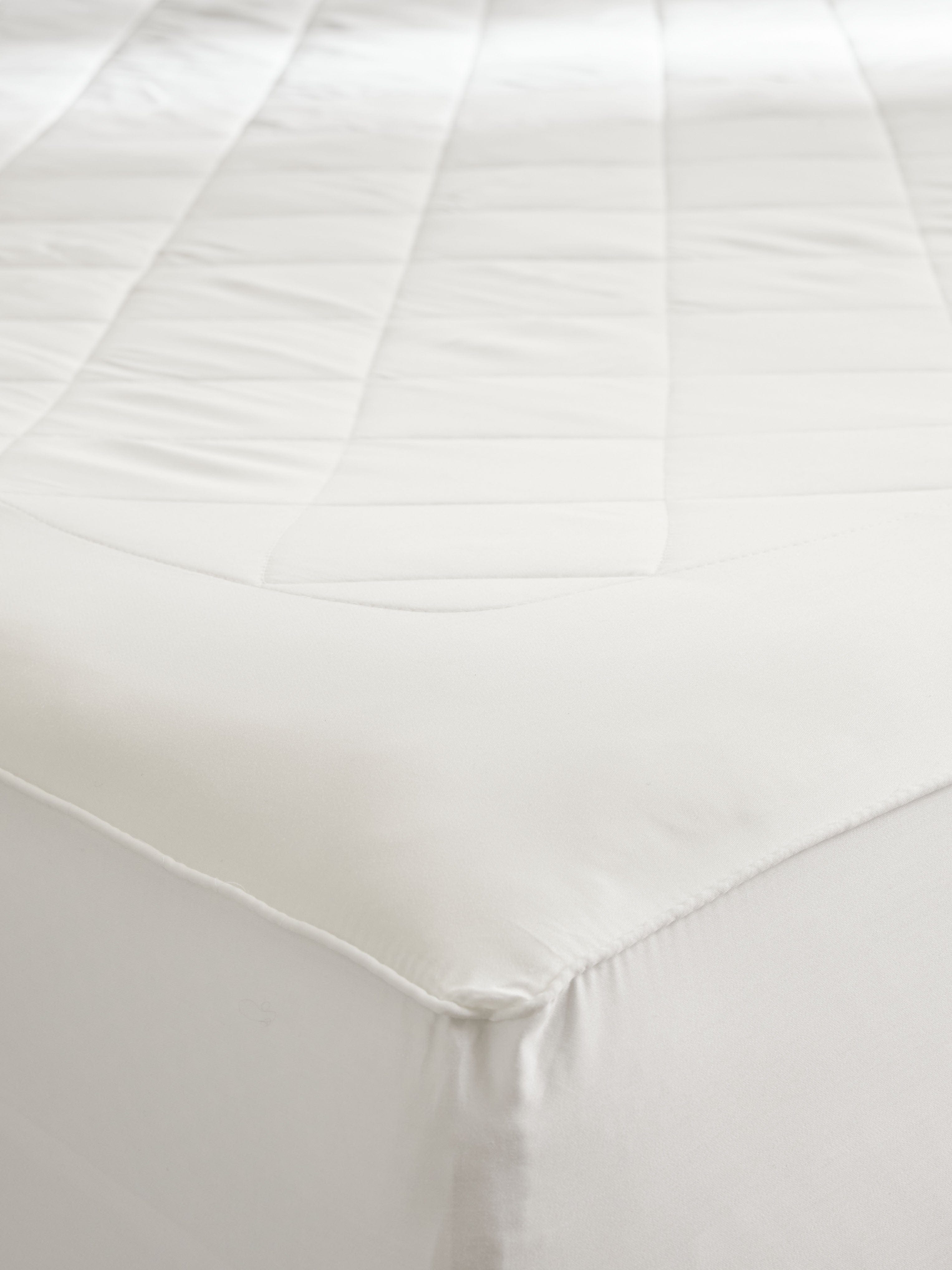 Detail of the mattress pad fitted around a mattress corner, showing the quilted top, smooth border, and fabric skirt wrapping down the sides.
