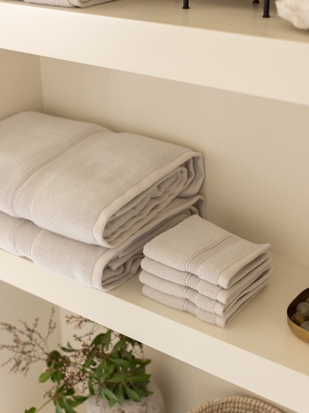 Ivory bath towels and washcloths arranged on a cream bathroom shelf, with folded edges facing outward and a leafy plant below.