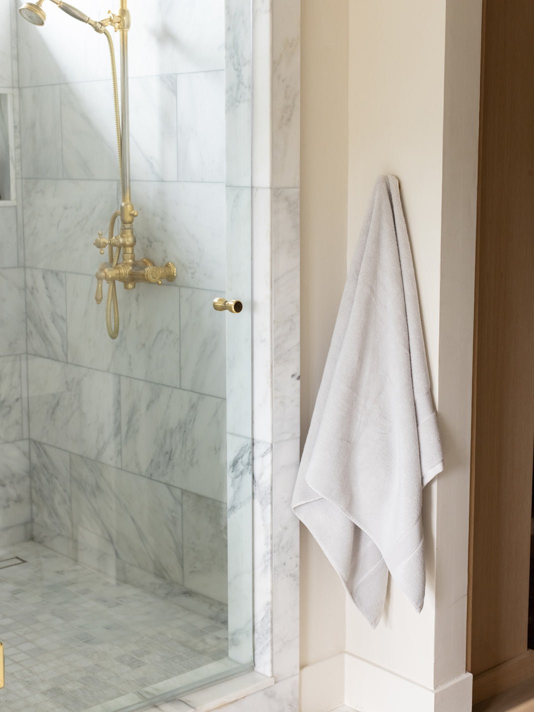 Ivory bath sheet hanging from a wall hook beside a glass-enclosed marble shower with gold fixtures, showing its length and folded drape.