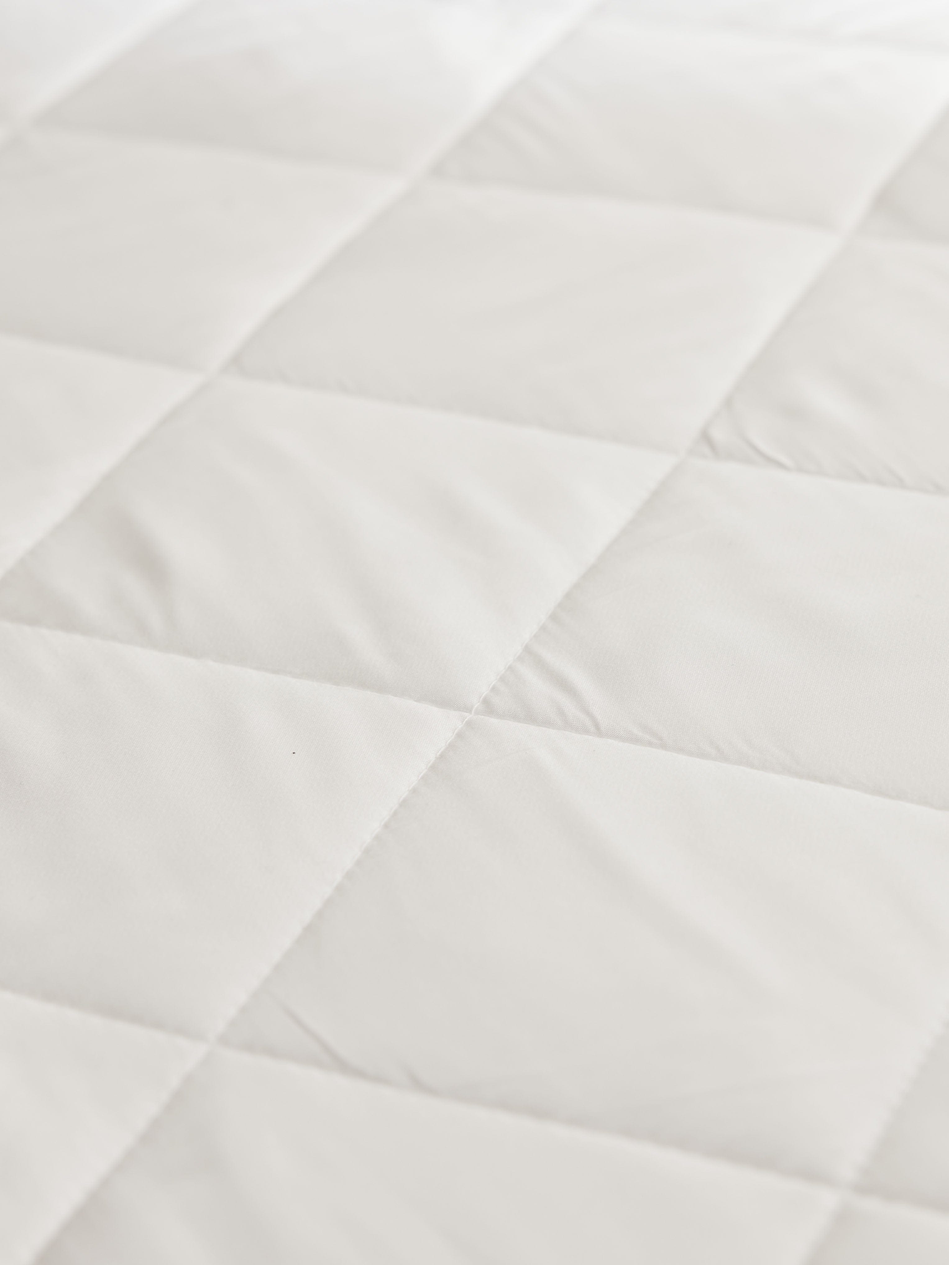 Close-up of the white mattress pad’s padded quilted sections, showing intersecting seams and subtle wrinkles in the fabric.
