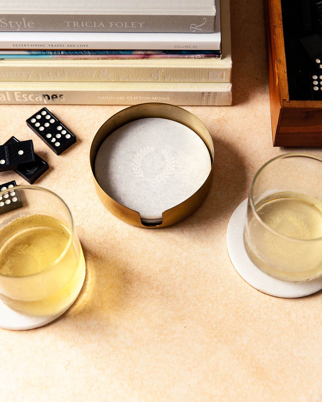 Overhead view of the marble coaster set among cocktail glasses, black dominoes, and a stack of books on a warm neutral tabletop.