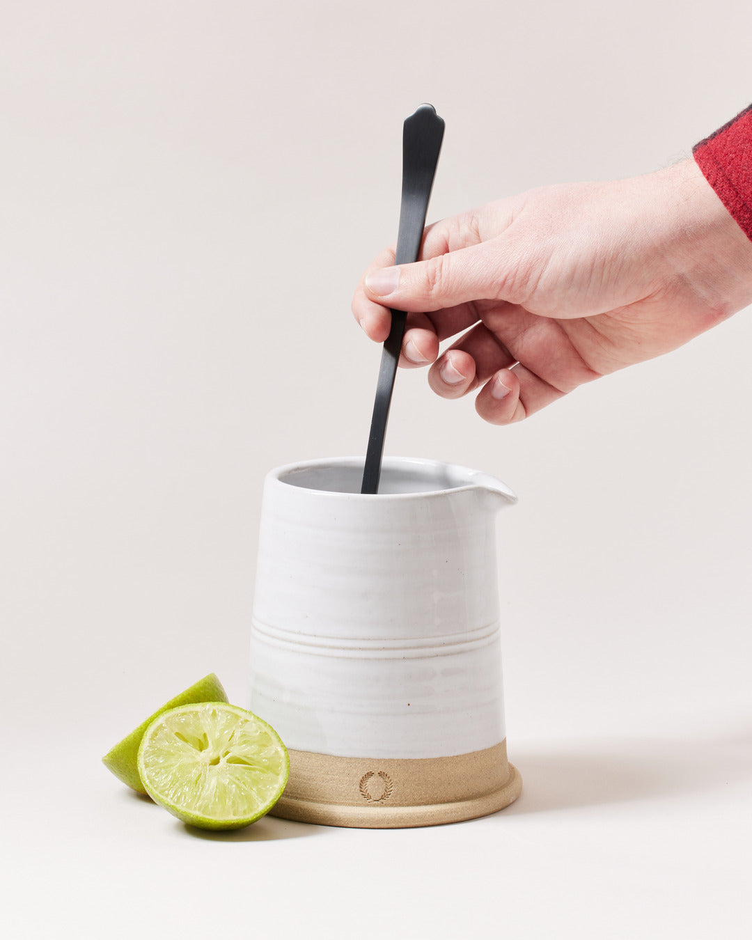 Hand stirring inside a white ceramic mixing vessel with the long onyx bar spoon, with a halved lime nearby.