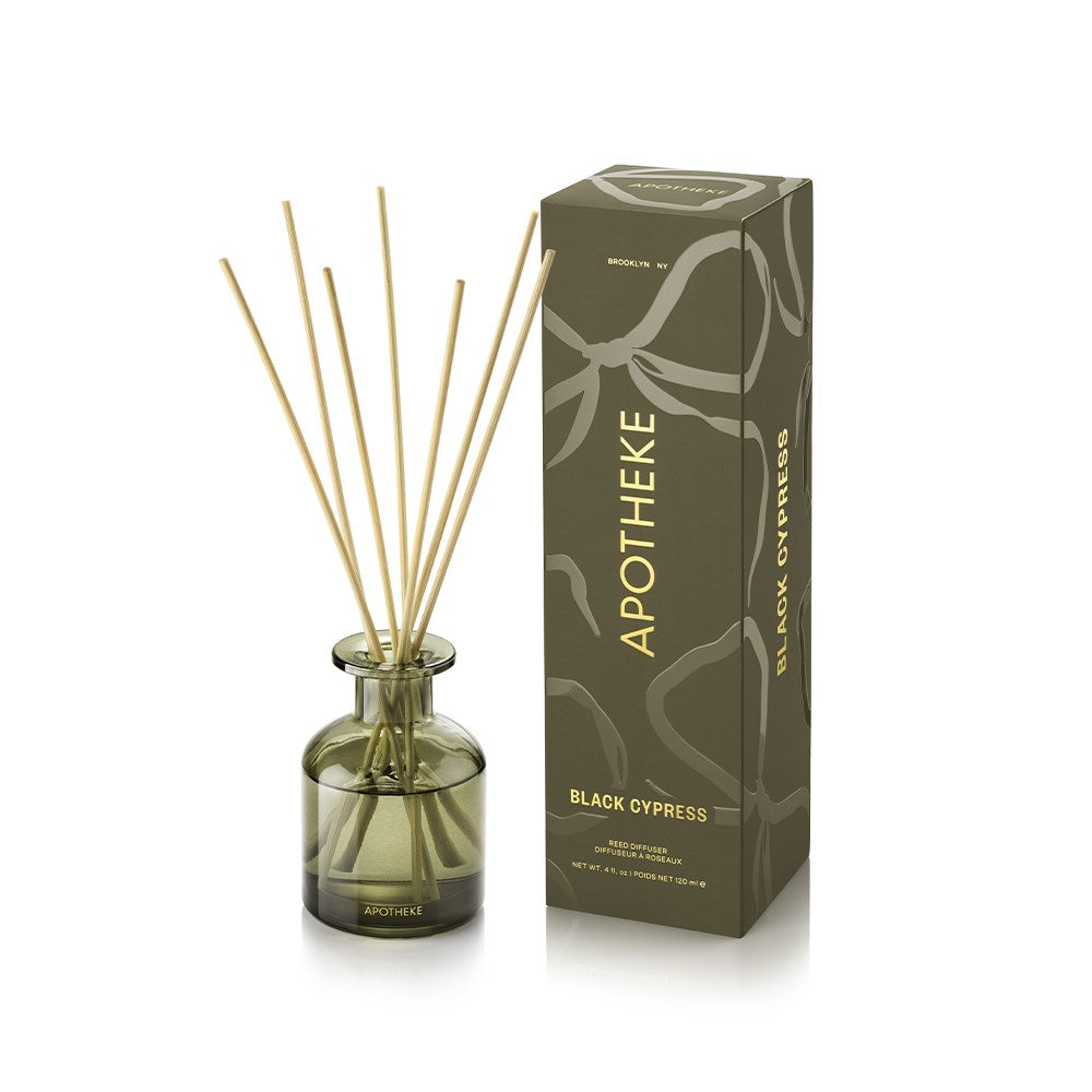 APOTHEKE Black Cypress Mini Reed Diffuser in an olive-green glass bottle with fanned natural-colored reeds, beside a tall olive patterned box with gold lettering on a white background.