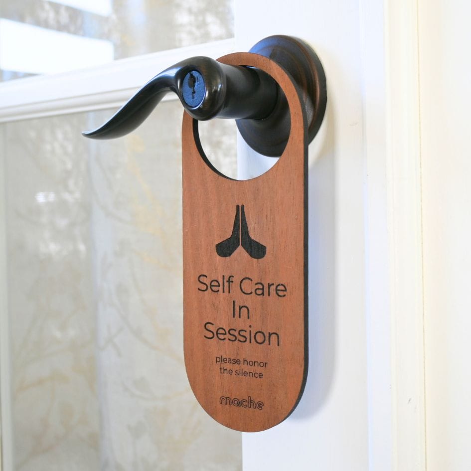 Dark walnut-colored wood door hanger on a black lever handle, printed with a meditation symbol and the message “Self Care in Session—please honor the silence.”