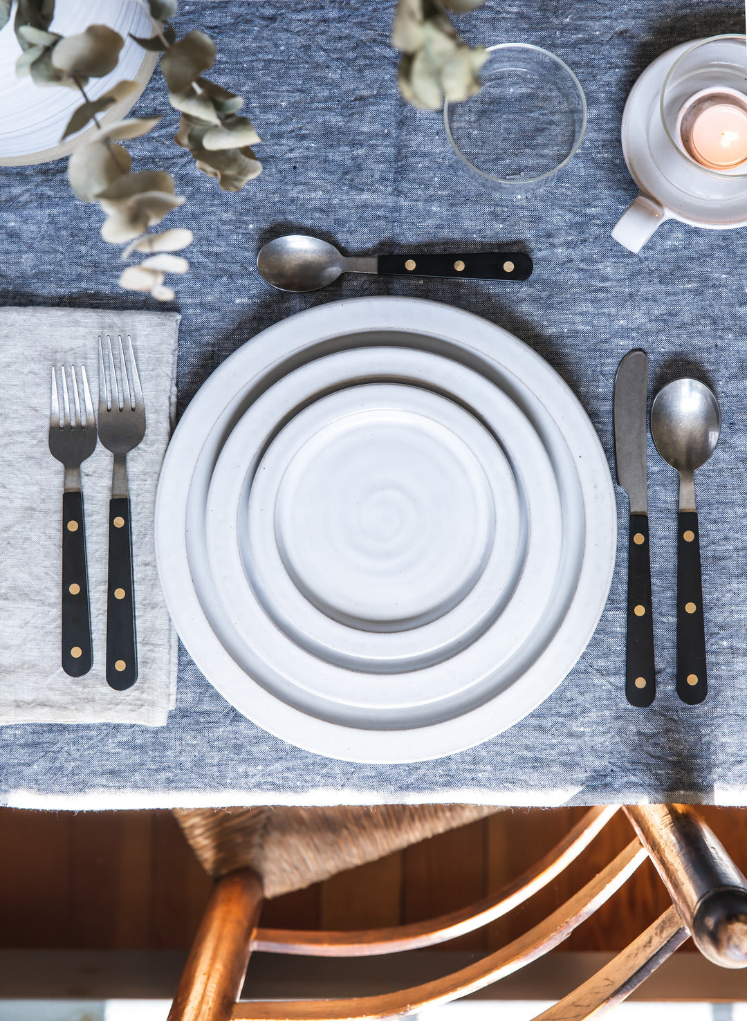 Black-handled Heritage Cutlery arranged around stacked white ceramic plates on a blue-gray linen tablecloth with glassware, greenery, and a small candle.