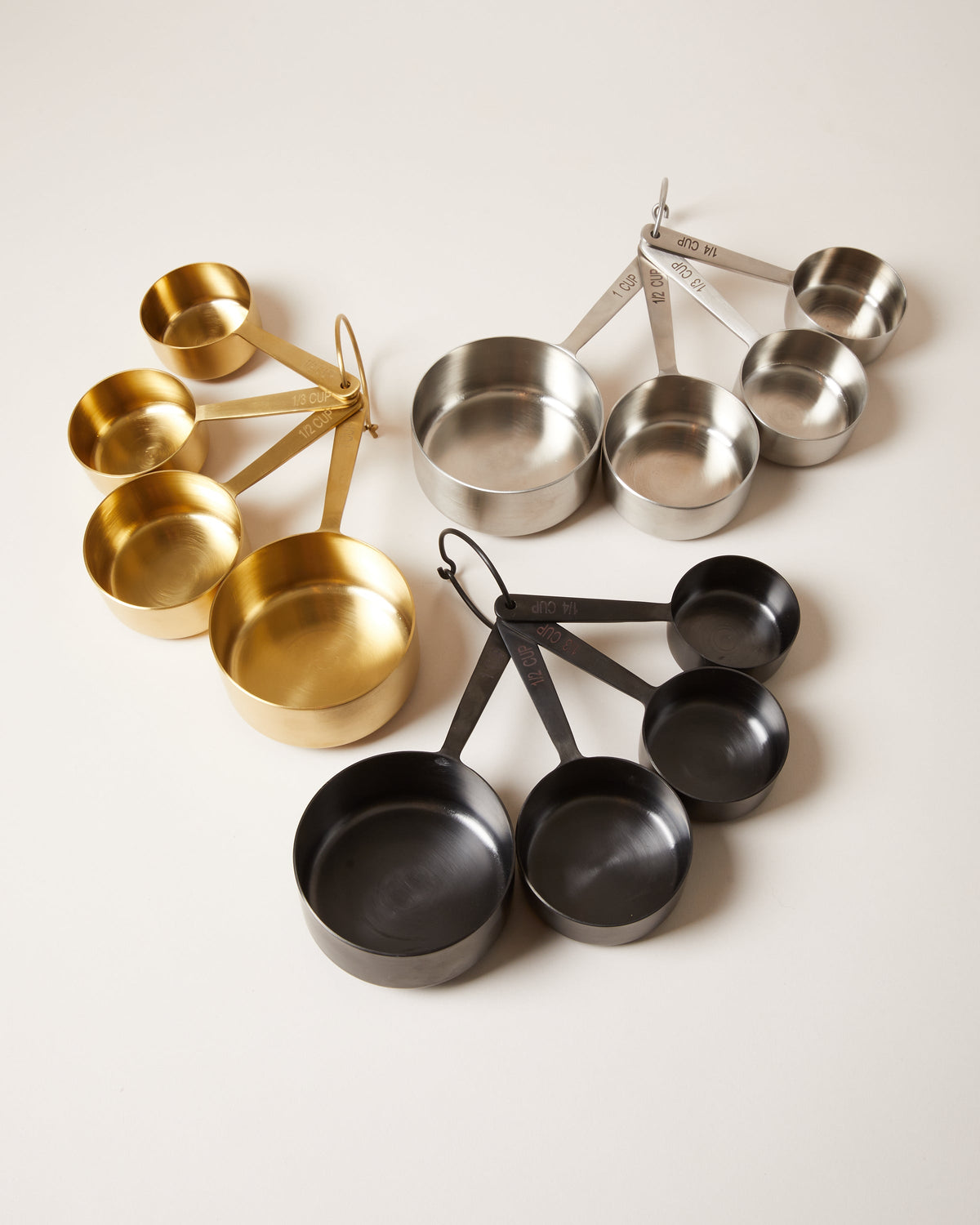 Three nested sets of four round metal measuring cups in brushed brass, stainless steel, and matte black finishes, each joined by a removable ring.