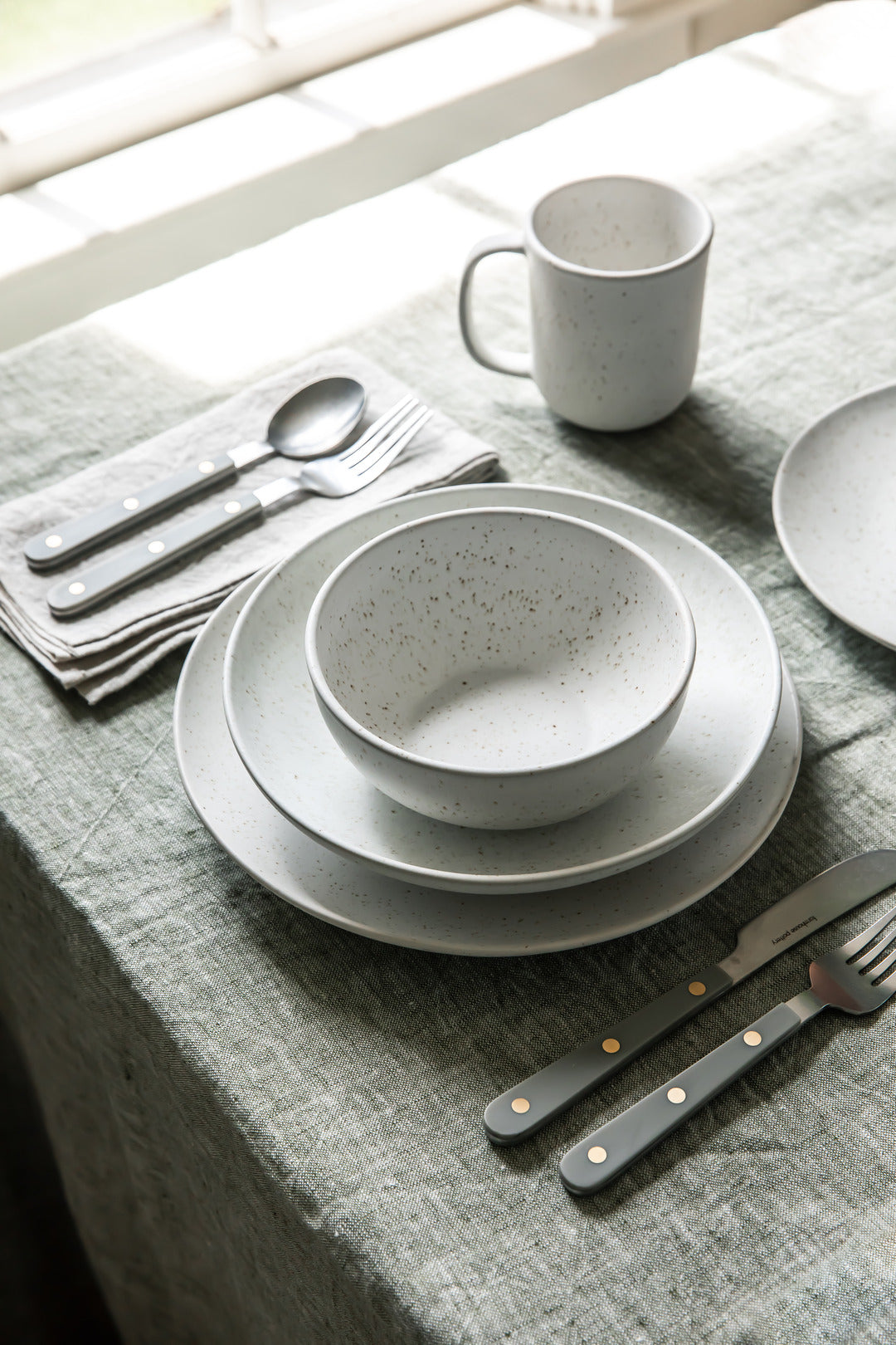 White speckled bowl and stacked plates arranged with a mug, flatware, and gray linen on a sunlit dining table.