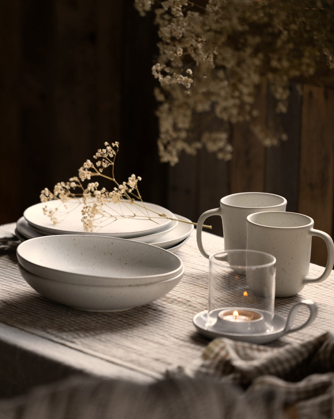 White Fieldstone bowls, plates, and mugs arranged with dried flowers and a glowing candle in a softly lit rustic tablescape.