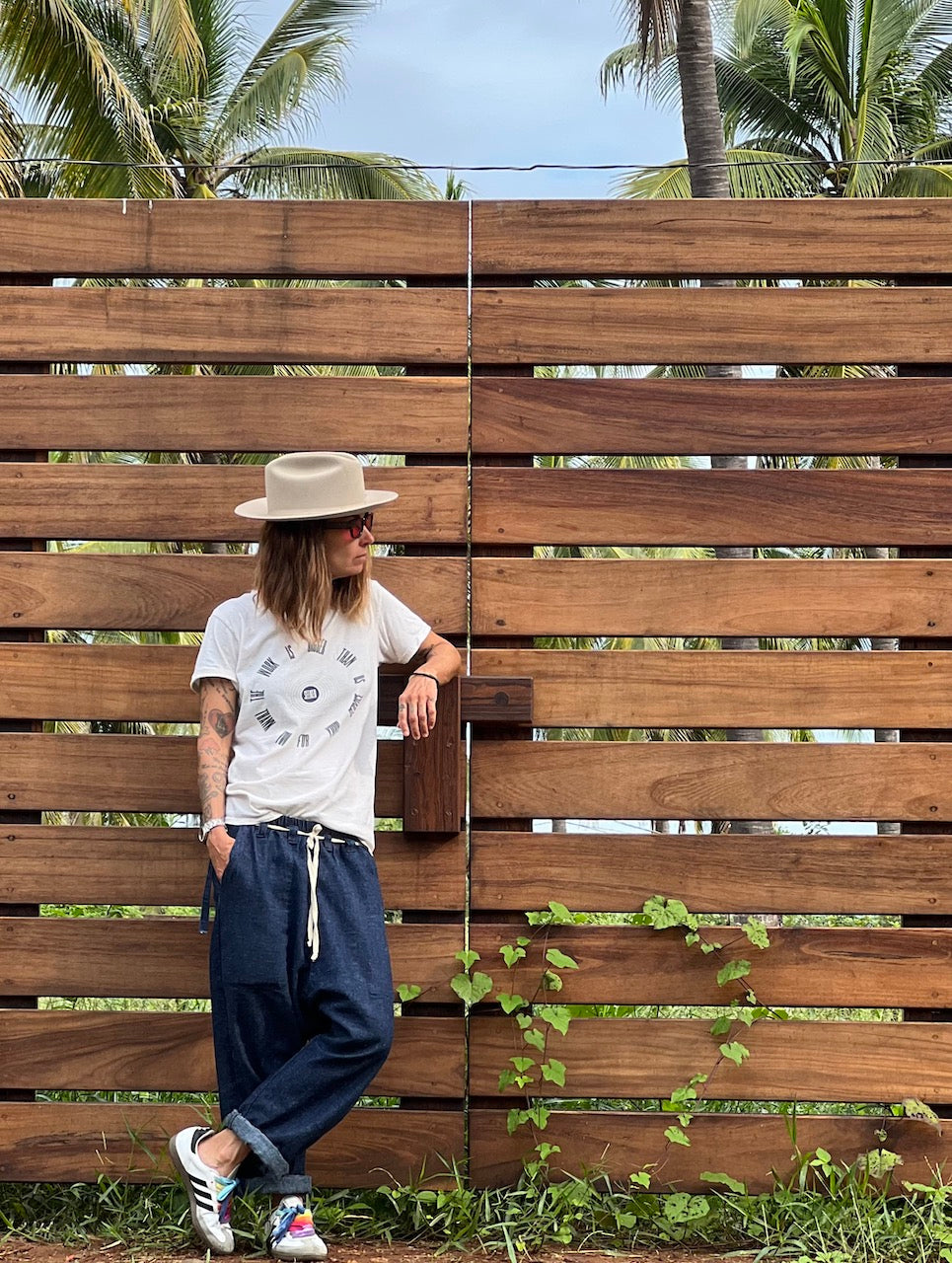 Model wearing a light gray Service Tee with a circular black graphic, loose navy drawstring pants, sneakers, and a wide-brim hat beside a wooden fence.