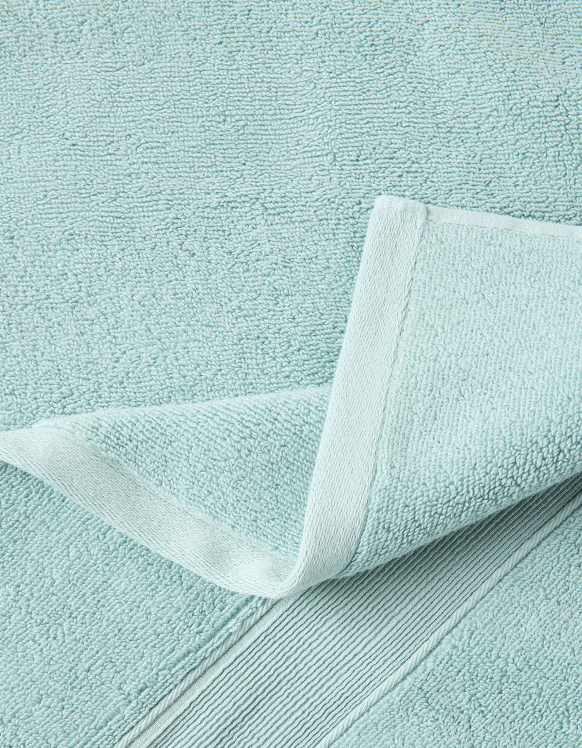 Close-up of a pale aqua towel with a corner folded back, revealing dense looped texture, a stitched edge, and a ribbed border.