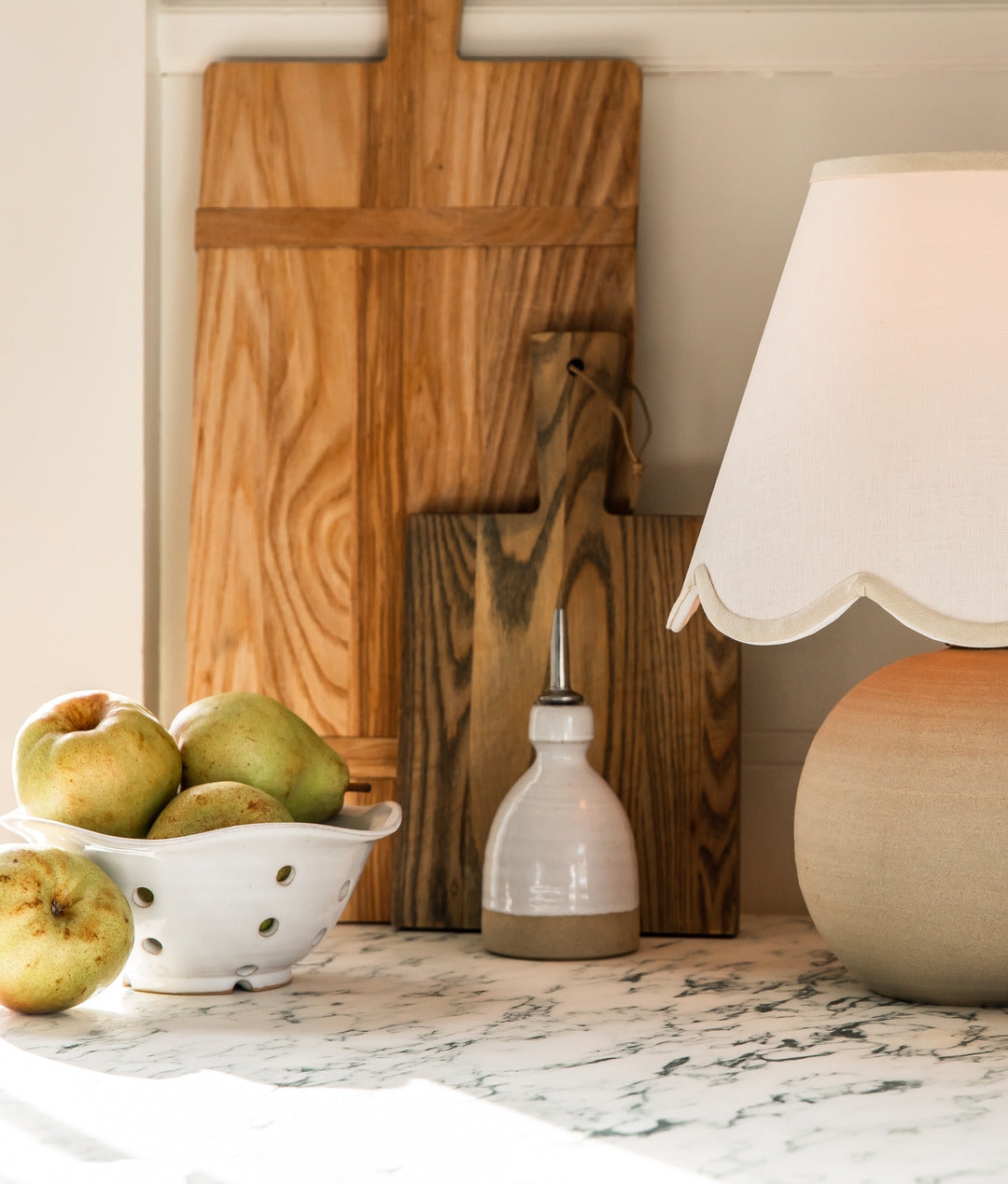 Tall wood cutting boards propped against a kitchen wall beside a ceramic lamp, a small bottle, and a white colander filled with pears.