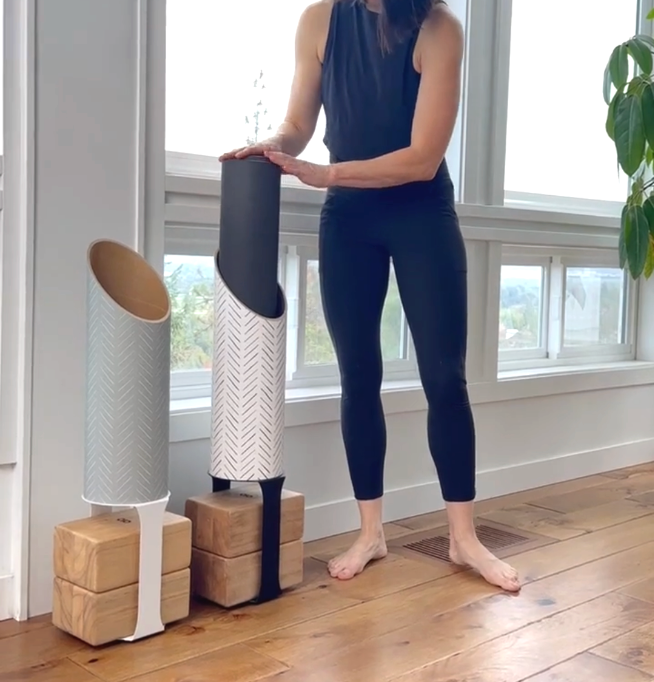 Woman in navy activewear placing a rolled black yoga mat into one of three Mache tubes displayed on wooden block stands.