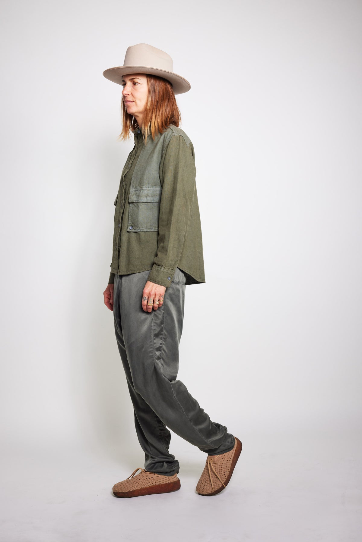 Side view of model walking in charcoal Souk pants, an olive jacket, and beige hat.