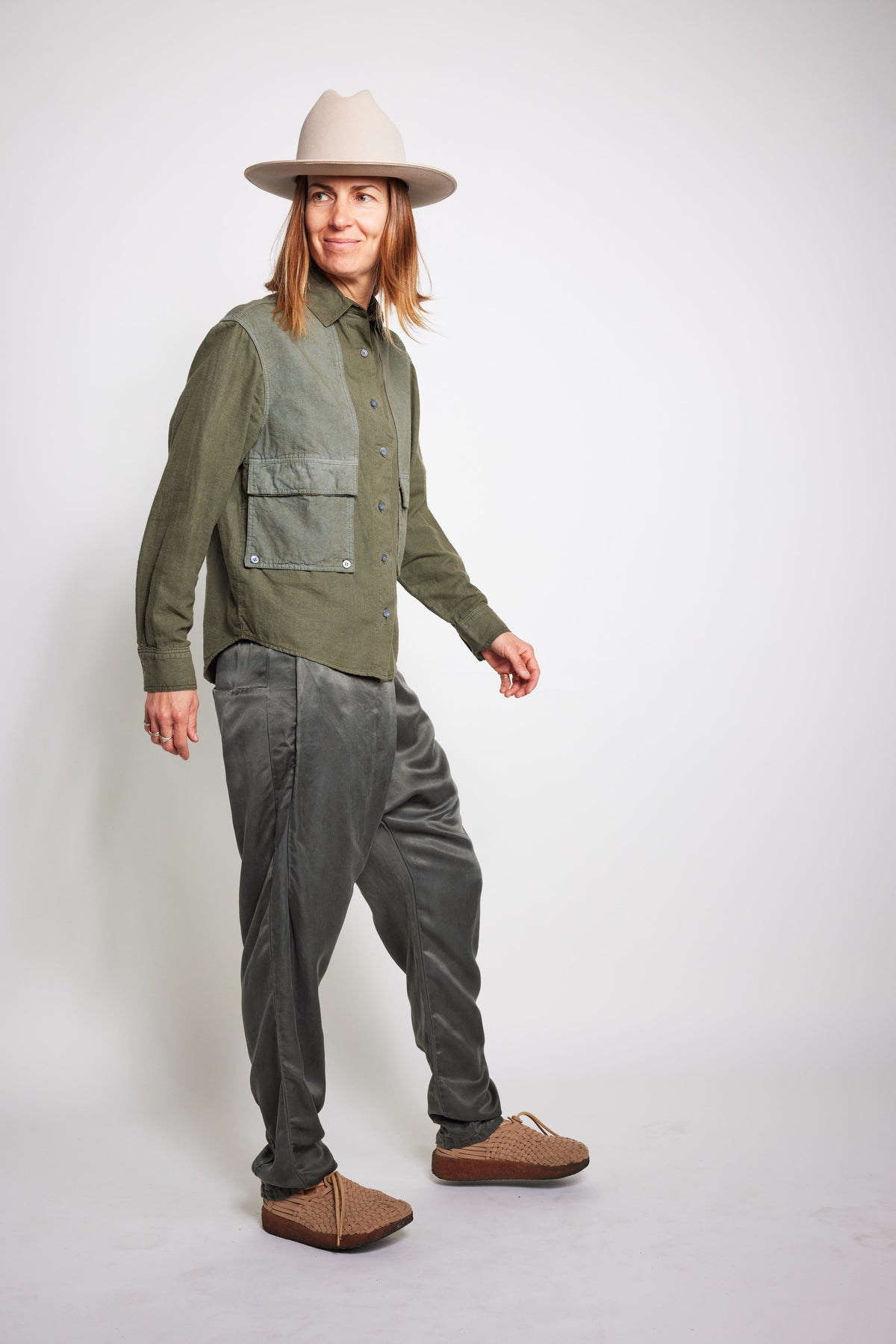 Model walking in an olive-green Trek Shirt with multiple utility pockets, gray pants, a beige hat, and brown shoes.
