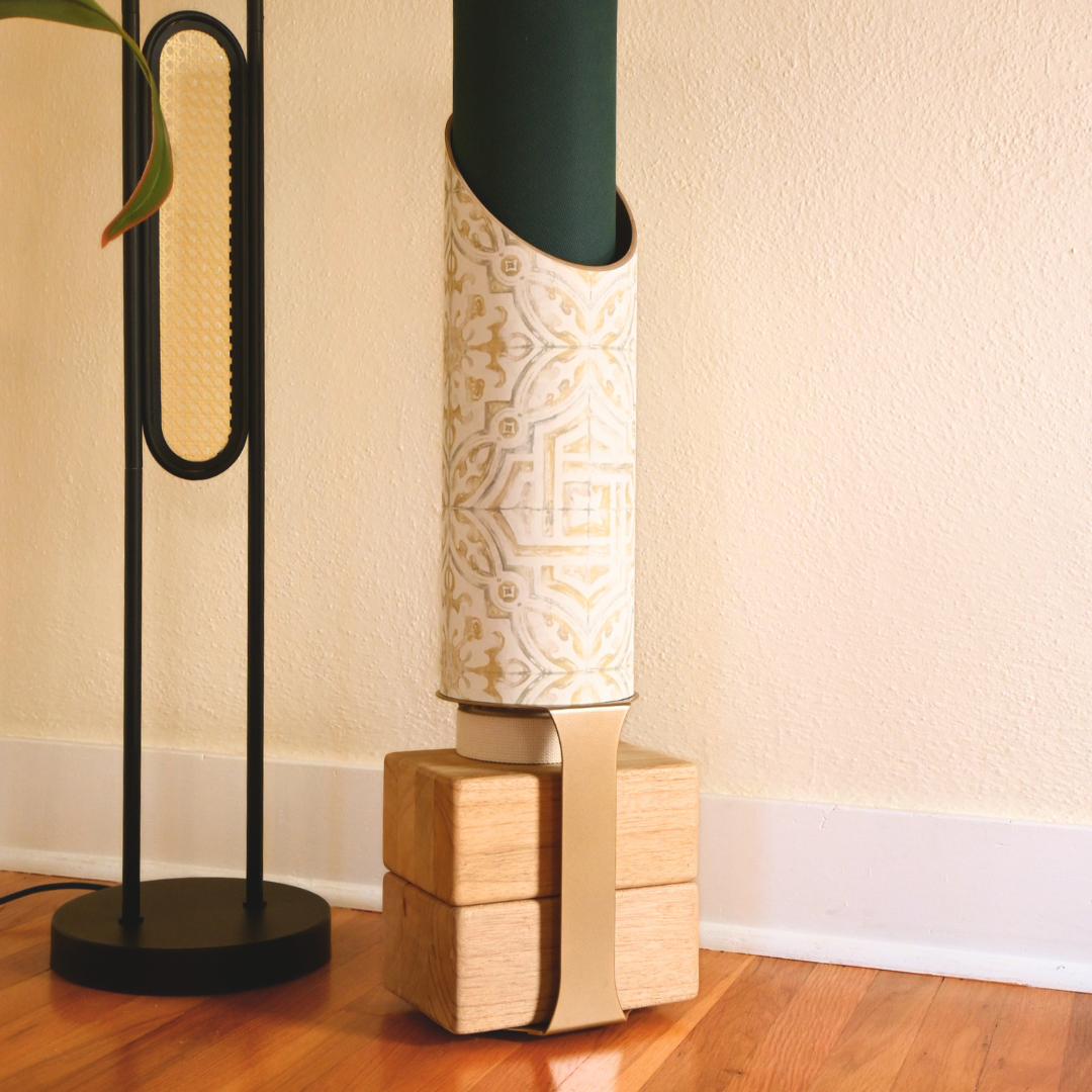 Star Crossed yoga mat tube displayed on its wooden base beside a black floor lamp and rolled dark green yoga mat.