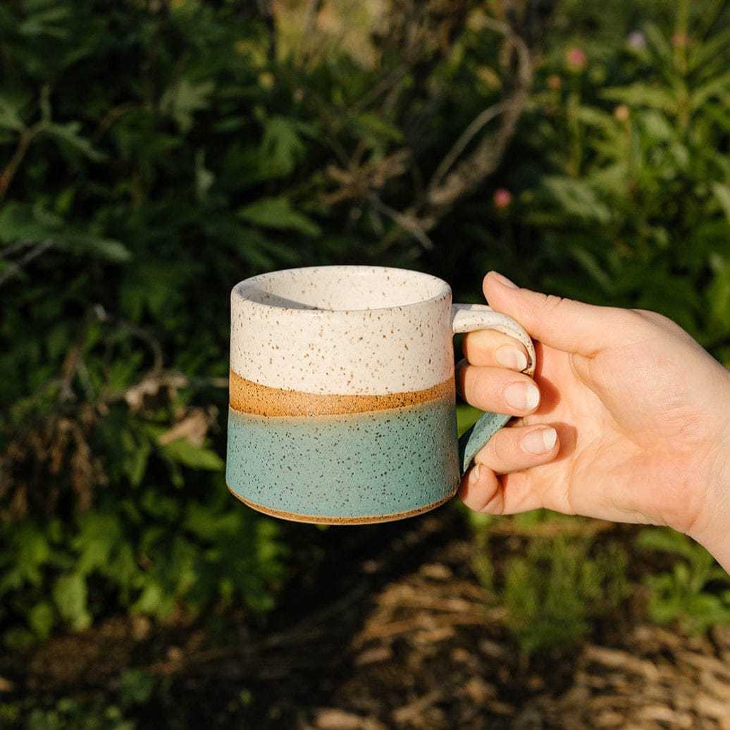 Hand holding a ceramic mug with a natural finish against a green outdoor background