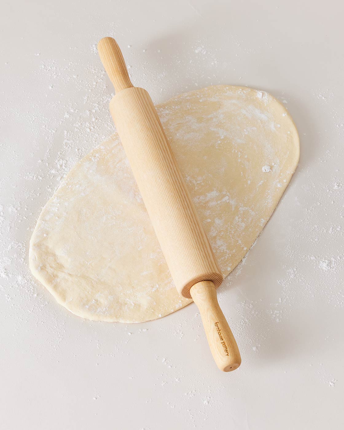 Natural ash rolling pin resting across pale, flour-dusted dough on a light neutral surface.