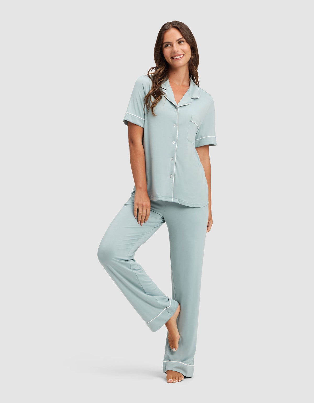 Model wearing a pale blue pajama set with a collared button-front top and matching relaxed pants, standing barefoot with one knee bent.