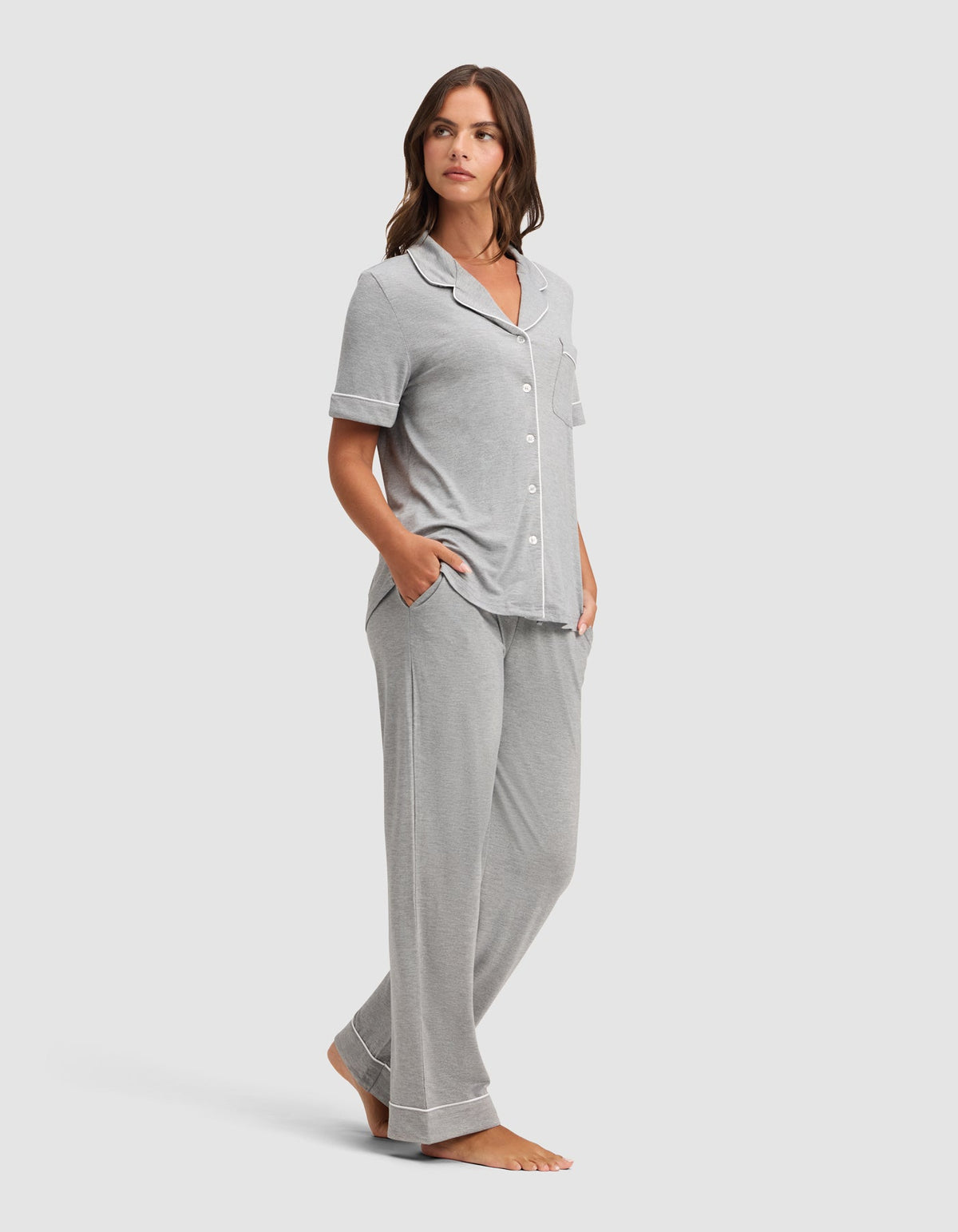 leg pants, with one hand in a pants pocket.
Model wearing a light gray pajama set with white piping, a notched collar, button-front top, and matching full-length pants, with one hand in a pocket.