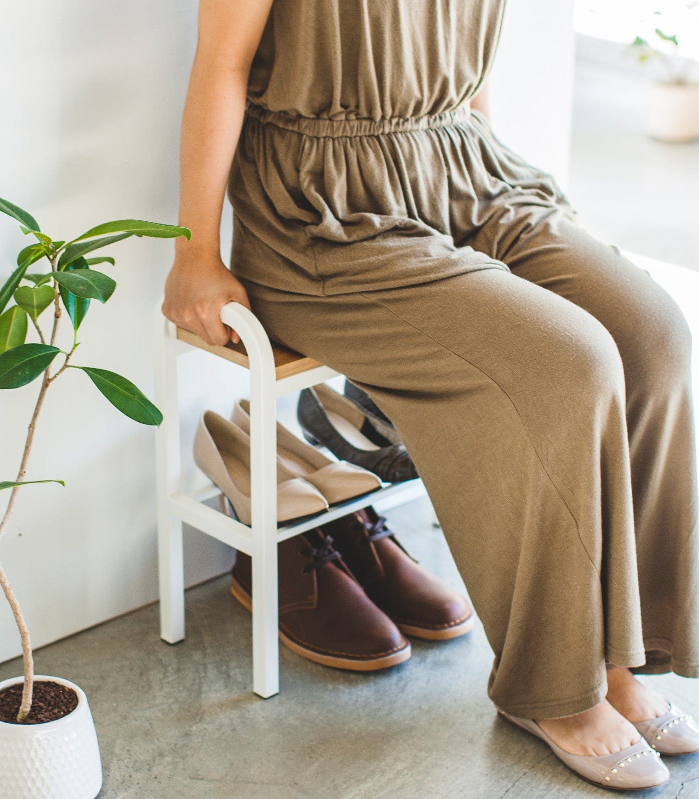 Person wearing a brown jumpsuit sitting on a chair with shoes underneath, in a room with a plant.
