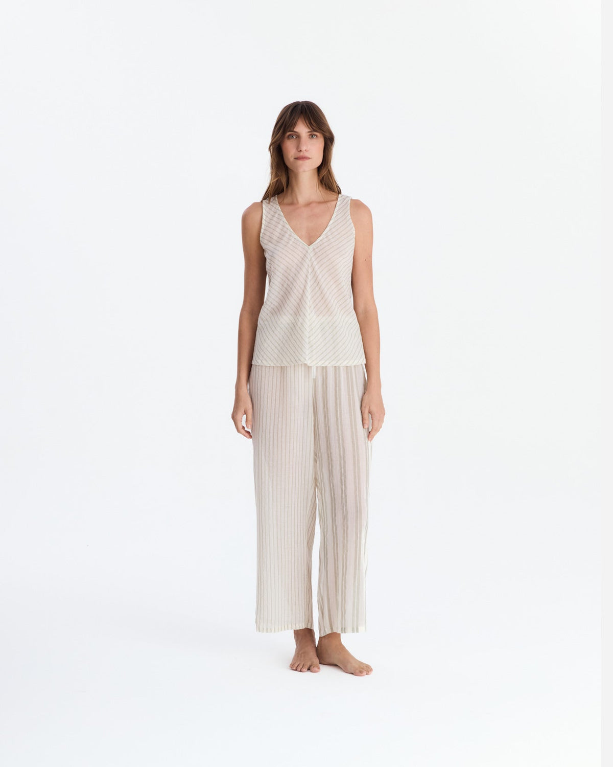 Front view of a barefoot model wearing cream and beige striped pajamas, with a sleeveless V-neck top featuring diagonal stripes and relaxed ankle-length pants with vertical stripes.