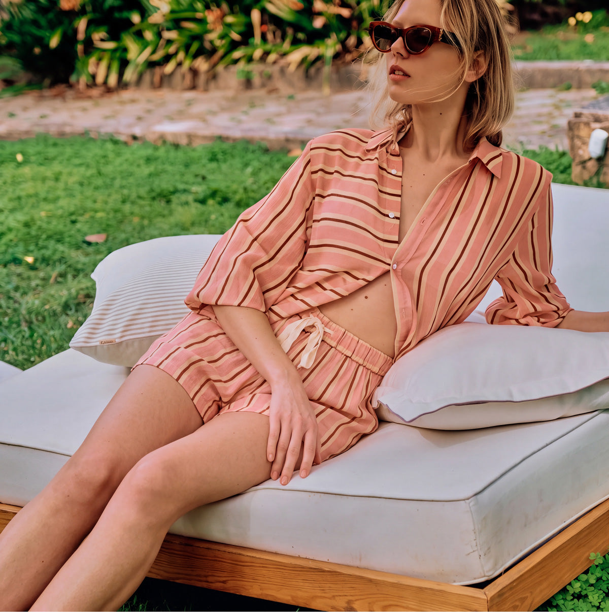 Model reclining on a cushioned outdoor lounge in coral striped pajamas, with the shirt partly open, sleeves pushed up, and sunglasses on.