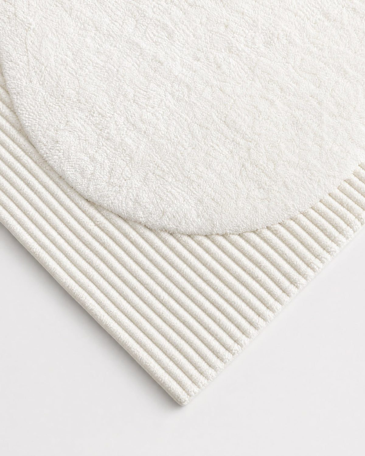 Close-up of the ivory bath rug’s corner, showing the curved edge of its plush oval center, parallel raised ribs, and neatly finished outer edge.
