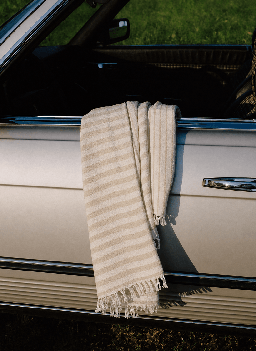 Beige and cream striped blanket draped over the open window of a light-colored car, with fringed ends hanging against the door.