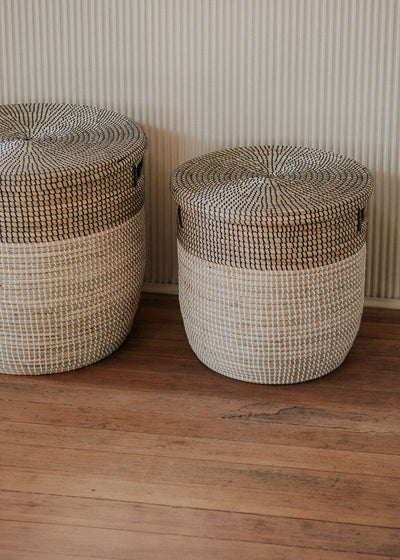 Pair of two-tone seagrass baskets arranged side by side against a softly striped wall.