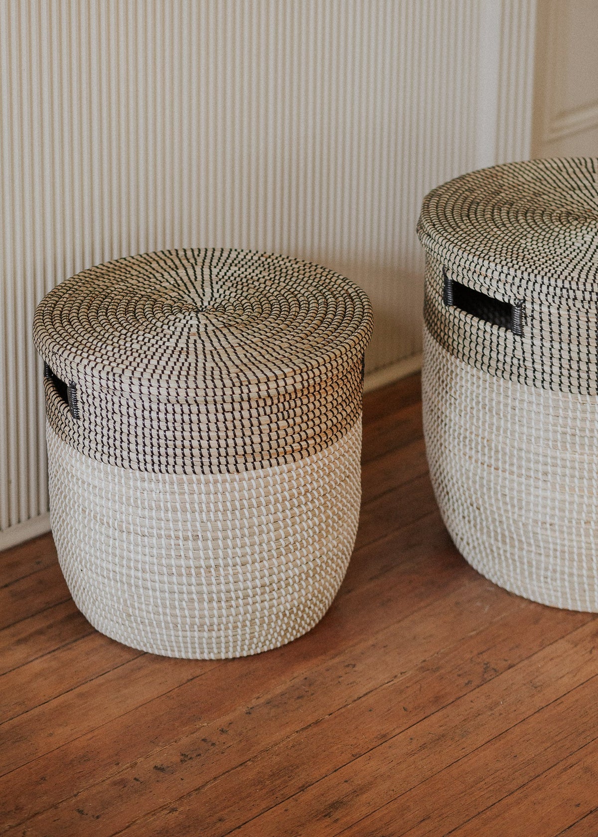 Two lidded Black Medina baskets styled on a wood floor in a paneled room.