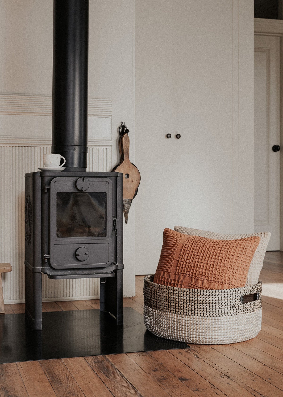 Black Medina floor basket holding a rust-colored cushion beside a black wood-burning stove.