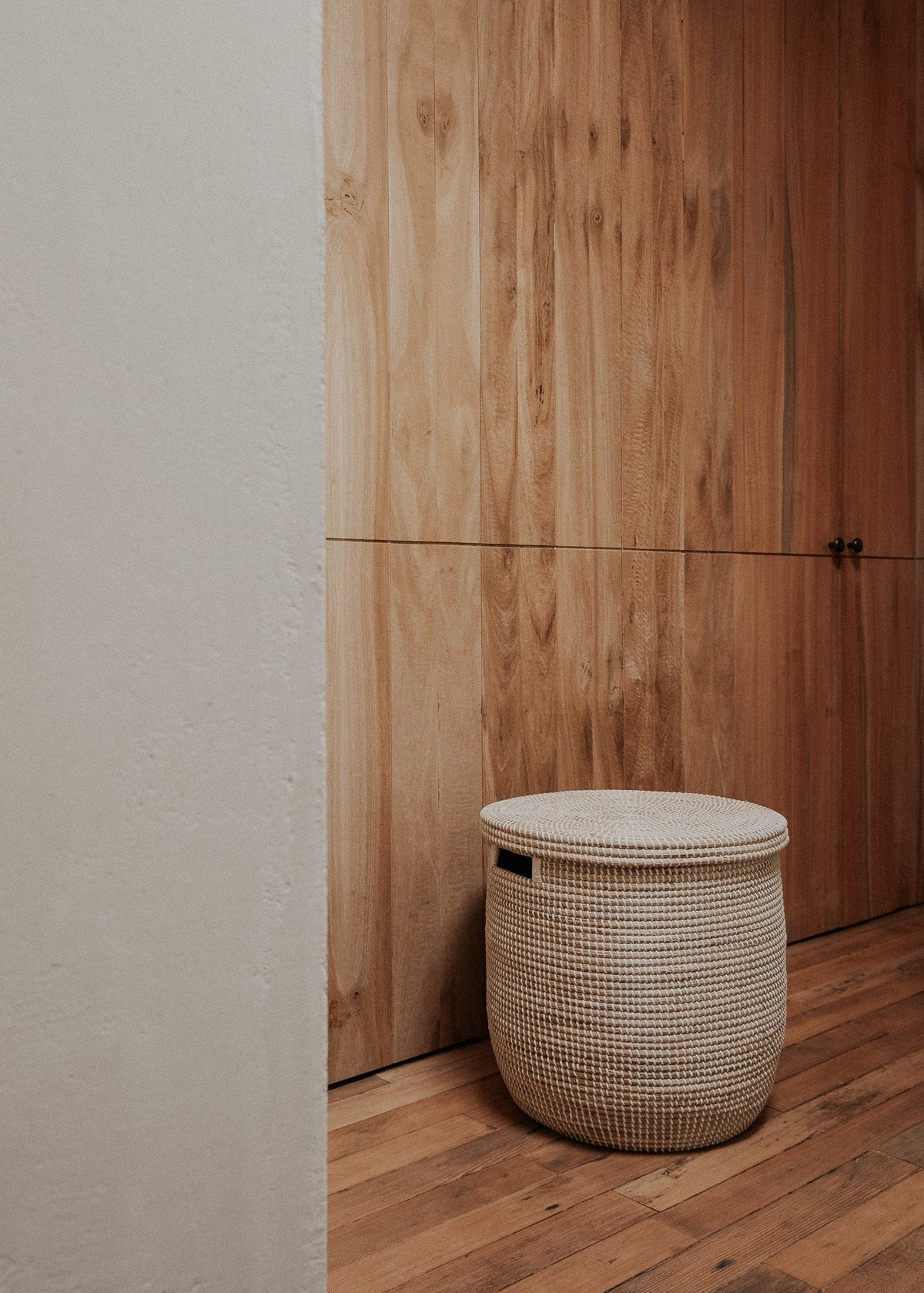 Large lidded Medina basket styled on a wood floor beside a tall natural wood cabinet.