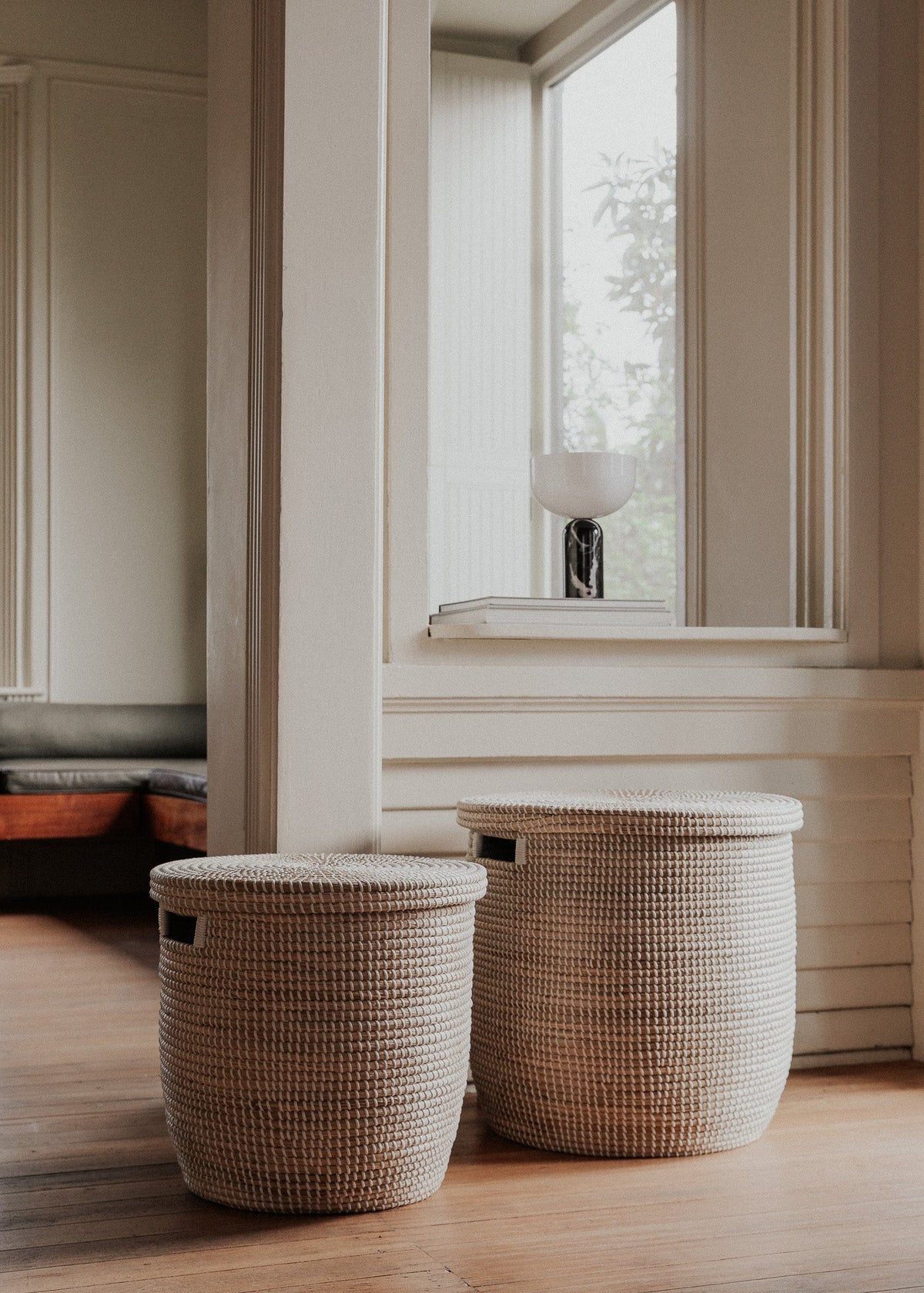 Two sizes of the lidded seagrass basket styled on a wood floor beside tall windows.