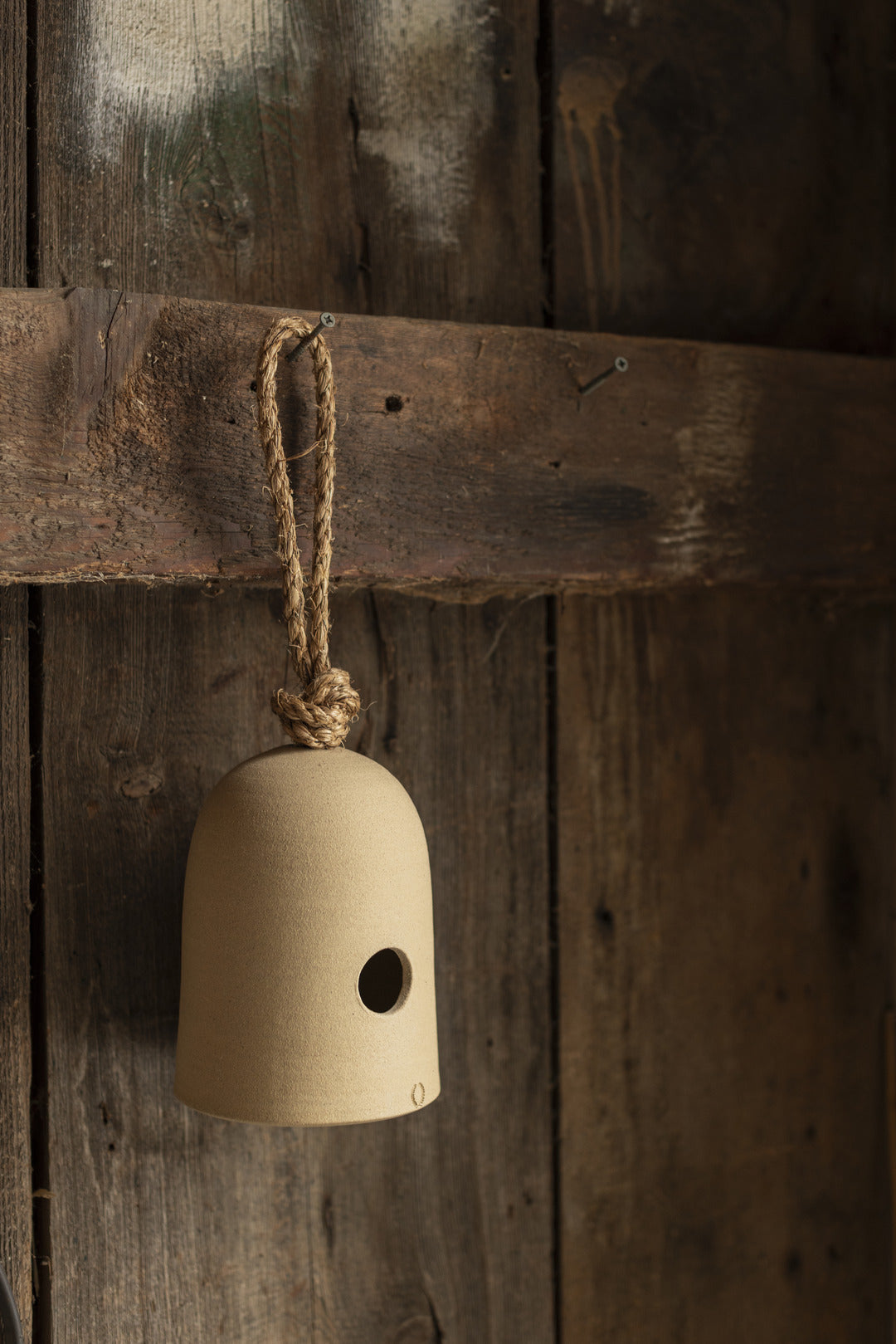 Cream ceramic bird shelter suspended by its rope loop against a weathered dark wood wall, showing the rounded dome and small circular entrance.