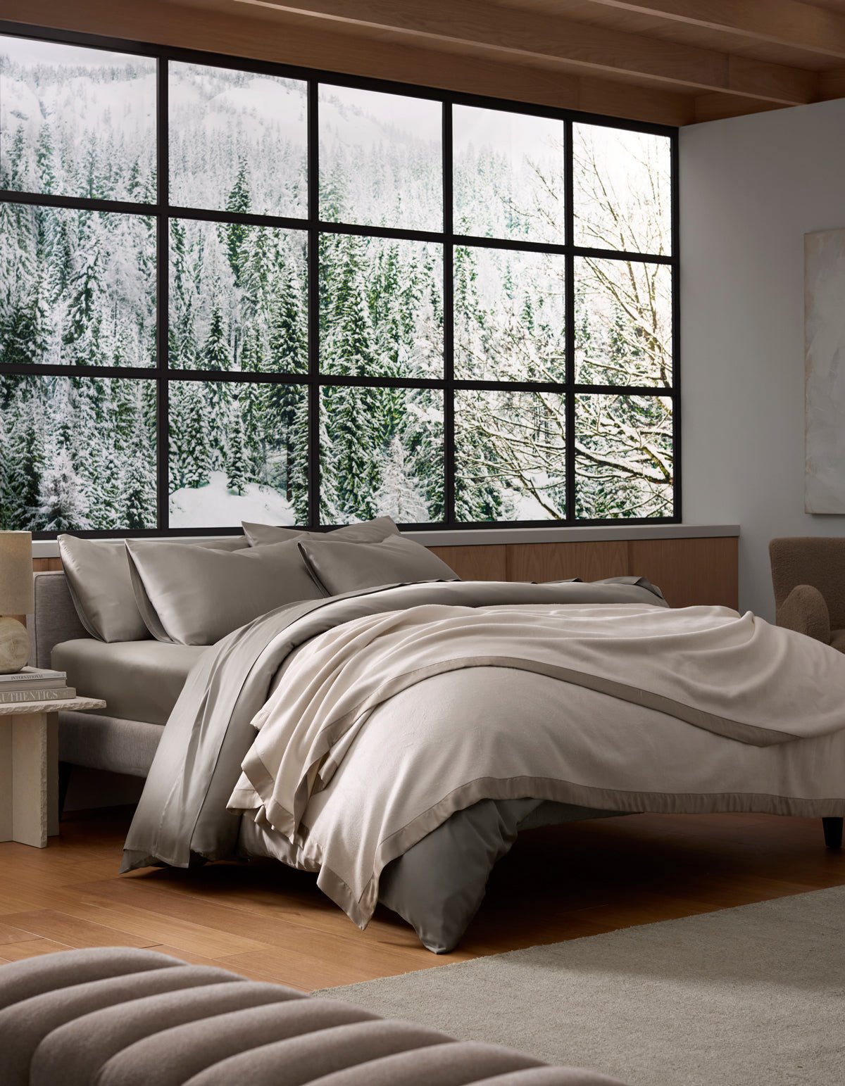 Ivory blanket loosely draped across a bed beneath large black-framed windows, with neutral pillows, a wooden floor, and greenery outside.