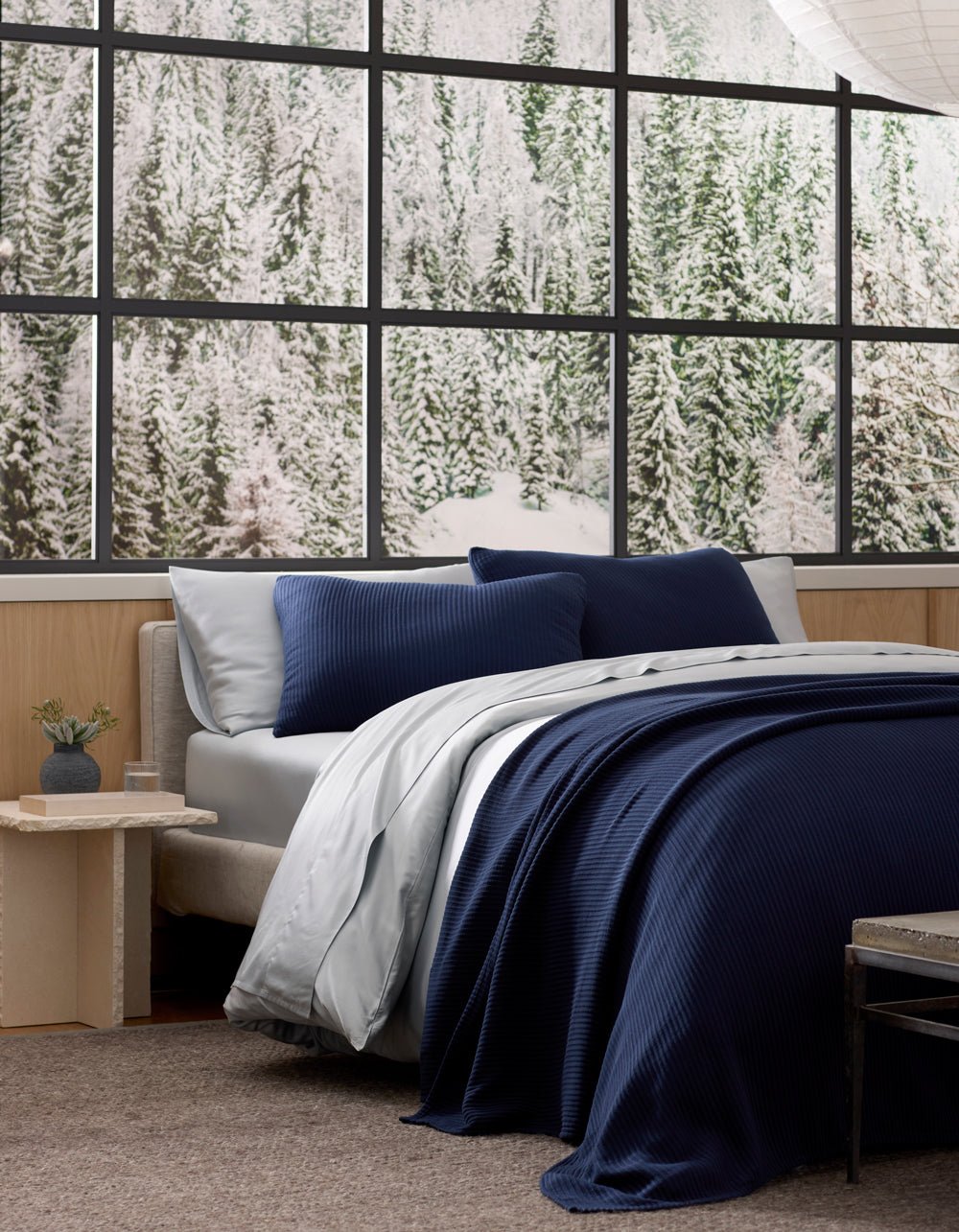 Navy coverlet draped over a bed with white bedding and navy pillows, beside large black-framed windows overlooking greenery.