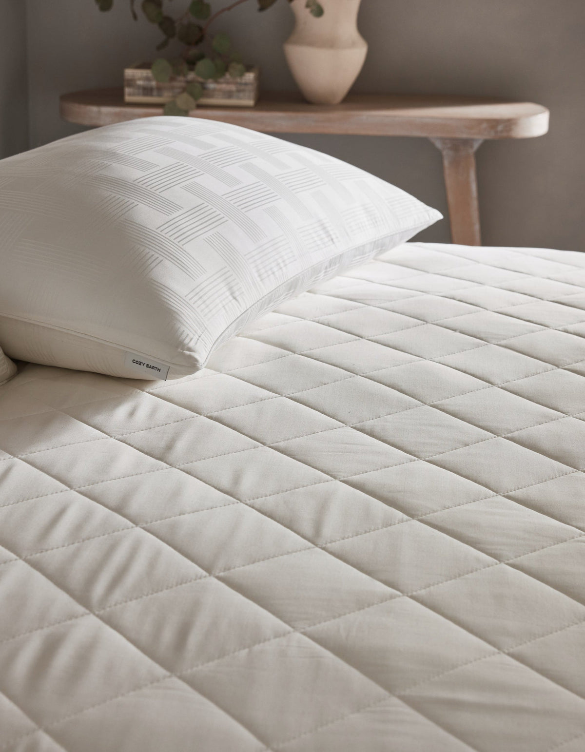 Angled close-up of the mattress pad’s quilted surface beneath a white pillow, with intersecting seams forming evenly spaced padded sections.