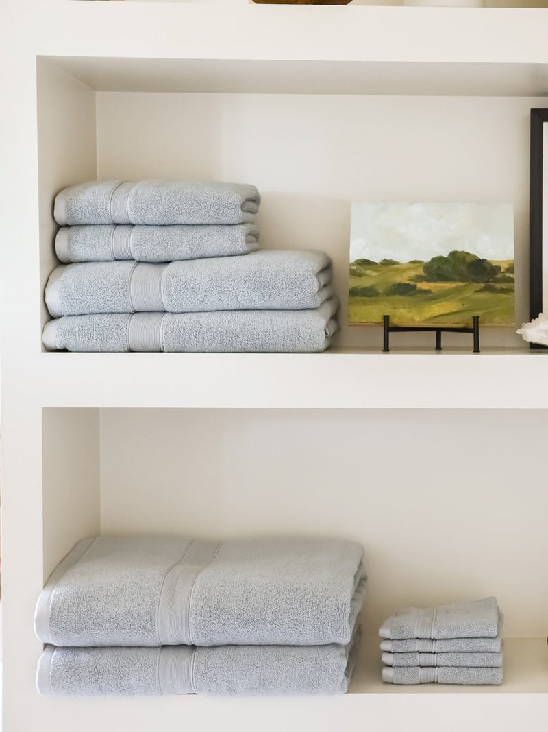 Pale blue towels displayed on cream shelves beside a landscape painting, with smaller towels above and larger towels and washcloths below.