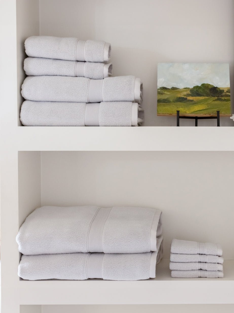Light gray towels arranged on two white shelves beside a small landscape painting, with larger folded towels and a separate washcloth stack below.