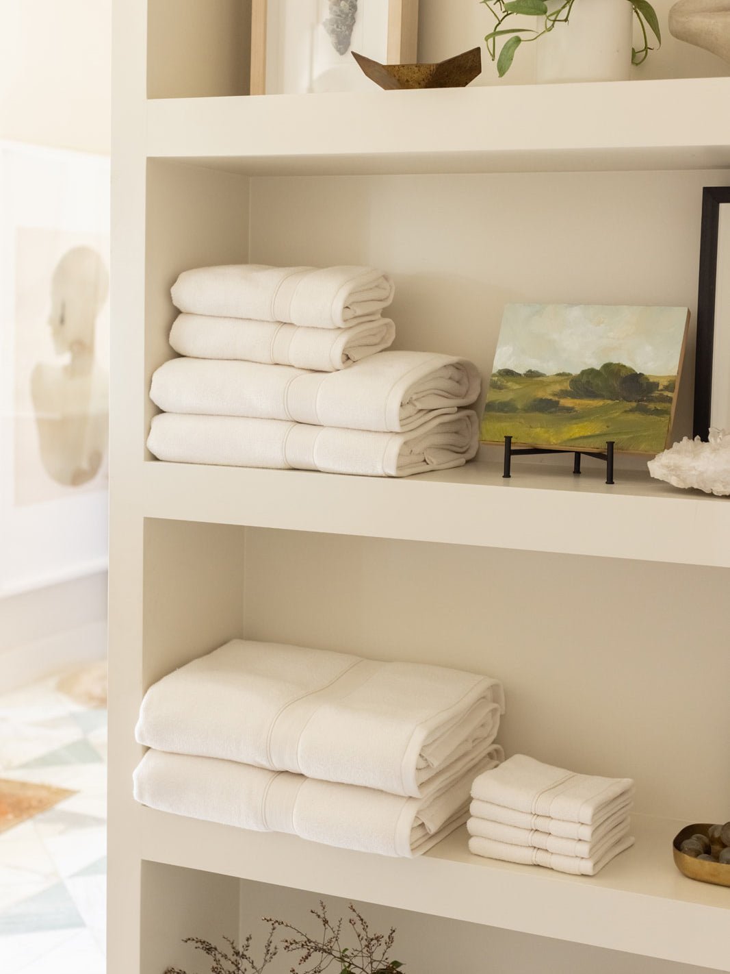 Ivory towels arranged on cream bathroom shelves beside a small landscape painting, with folded larger towels and a neat stack of washcloths on the lower shelf.