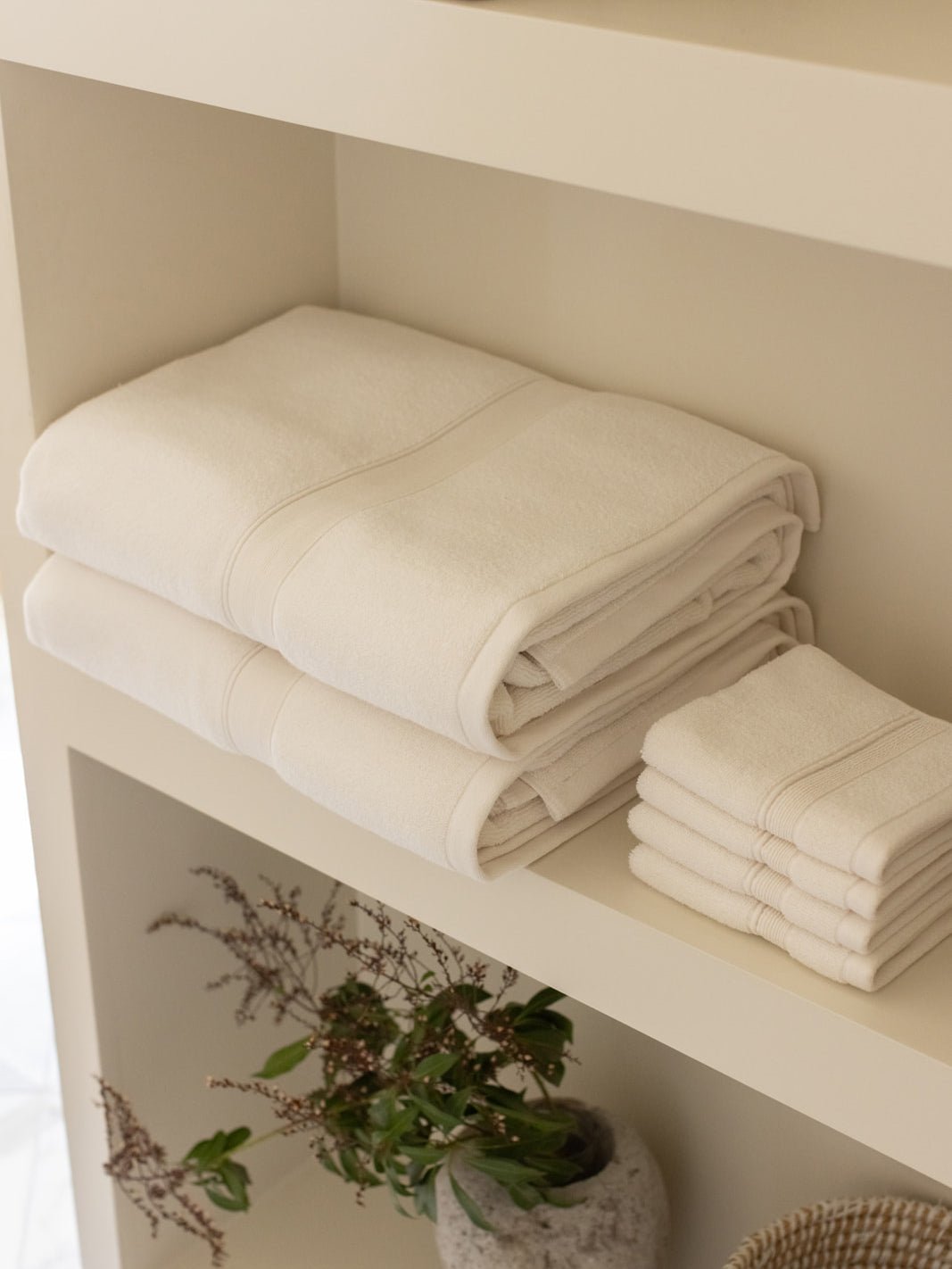 Ivory bath towels stacked on a cream bathroom shelf beside smaller folded towels, with a leafy potted plant below.