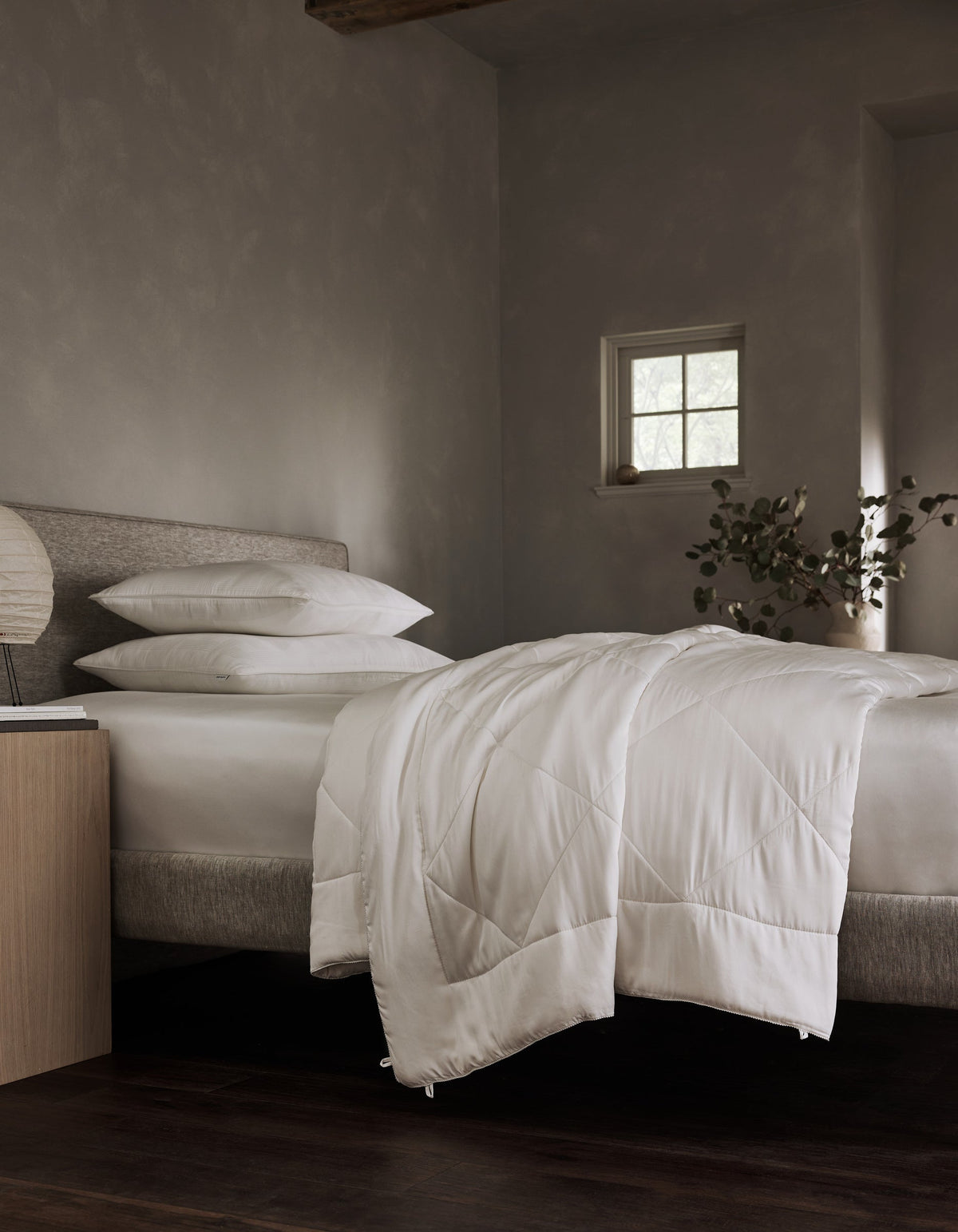 White quilted comforter hanging over the foot of a bed with layered white pillows, gray walls, and a small square window.